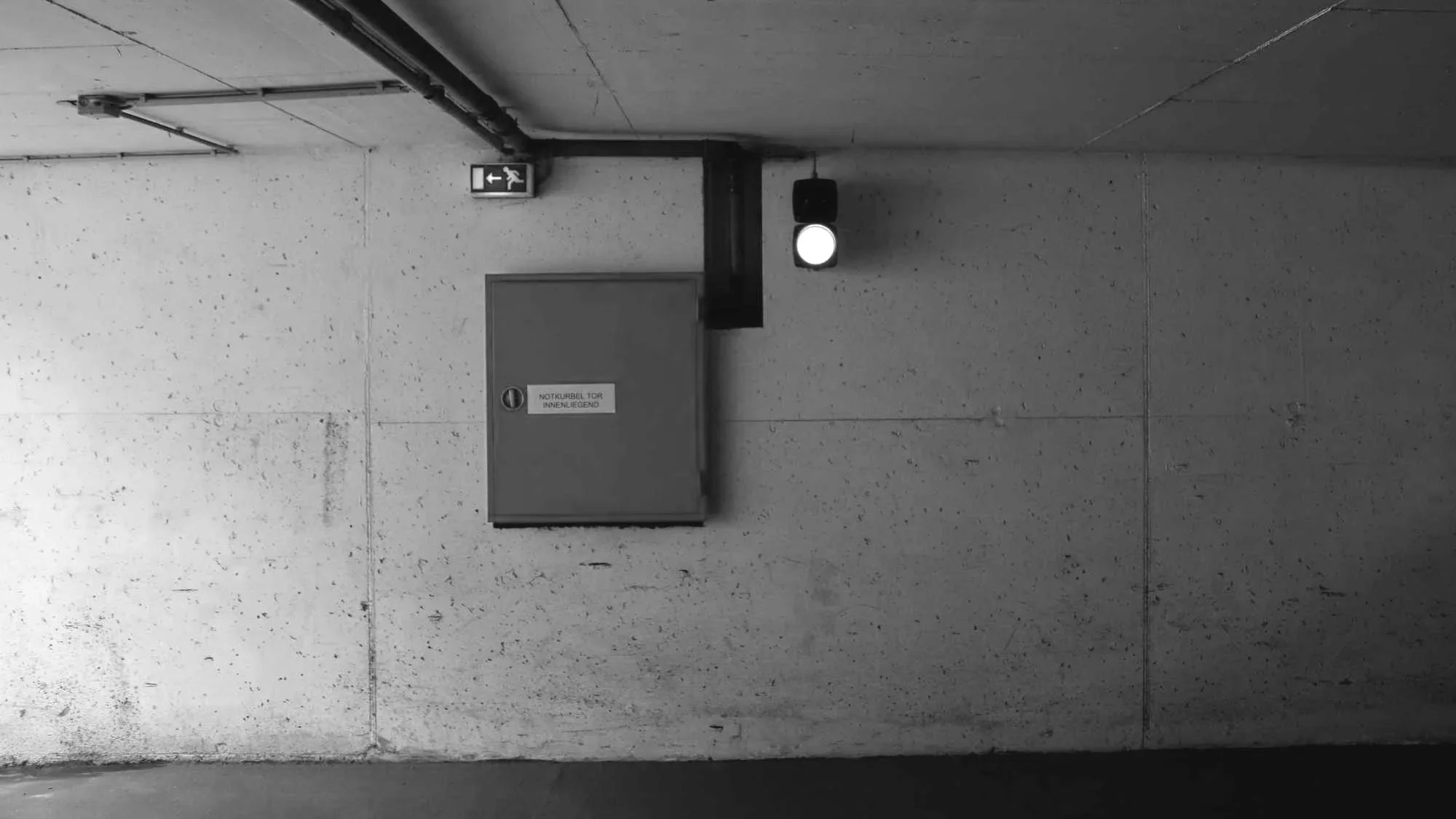 Ein beleuchteter grüner Signalhahn in einem Parkhaus. Auf der Wand befindet sich ein roter Feuerlöscherkasten mit der Beschriftung 'NOTAUSGANG TOR INNEN'. Über dem Feuerlöscher befindet sich ein kleines grünes Fluchtwegzeichen. Die Wand ist weiß mit einer rauen Oberfläche.