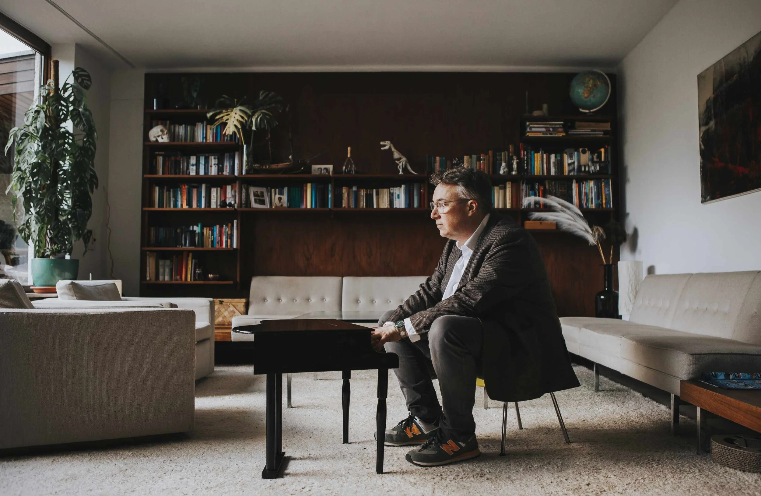 Ein Mann in einem Wohnzimmer sitzt auf einem schwarzen Stuhl vor einem kleinen schwarzen Spielzeug-Flügel, umgeben von weißen Sofas, mit Bücherregalen im Hintergrund und einer großen Zimmerpflanze auf der linken Seite.