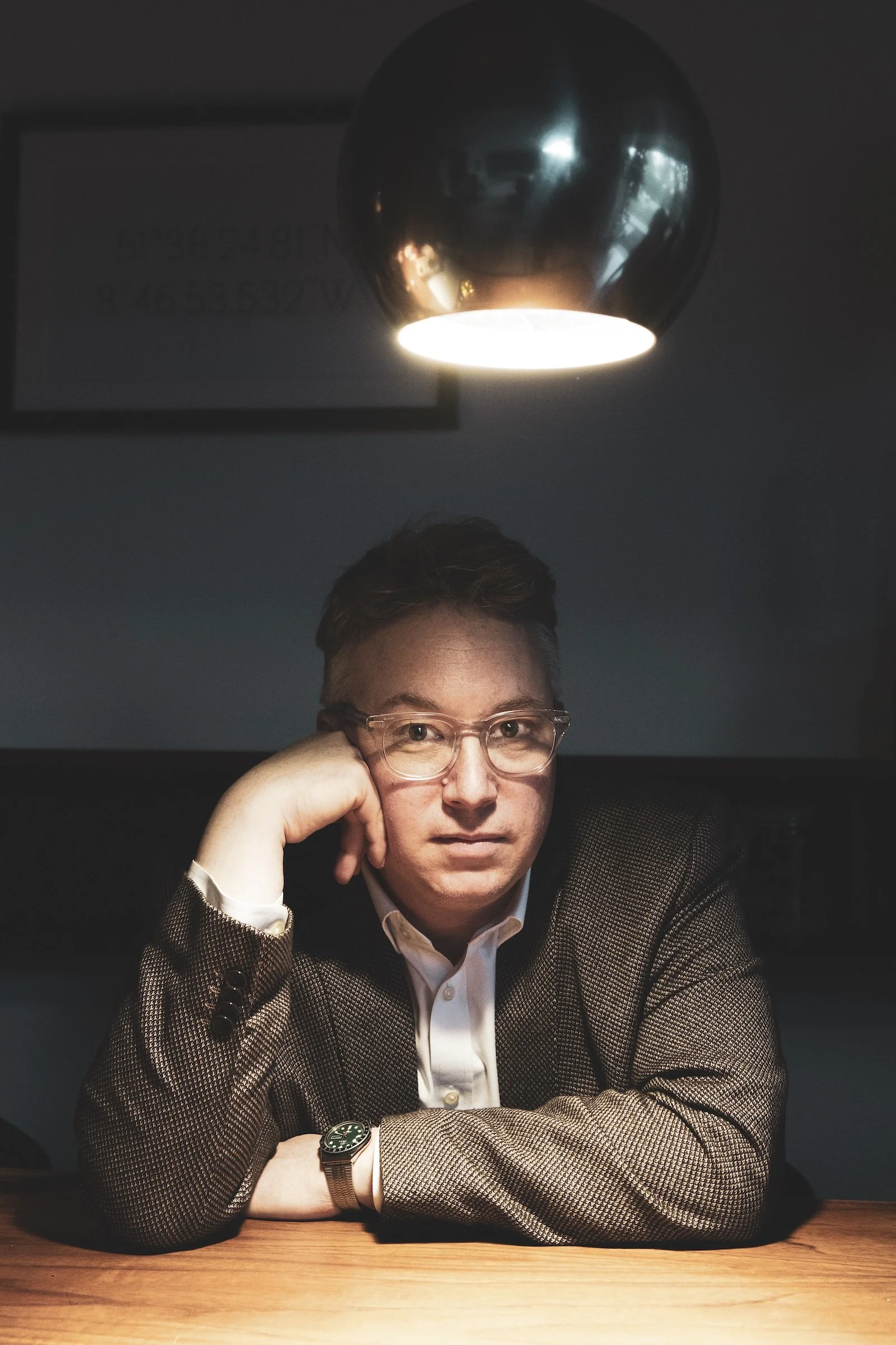 Junge Person mit Brille in einem dunklen Raum, sitzt an einem Tisch, trägt einen weißen Hemd und einen braunen Blazer, schaut direkt in die Kamera, unter einer modernen, silbernen, glänzenden Leuchte