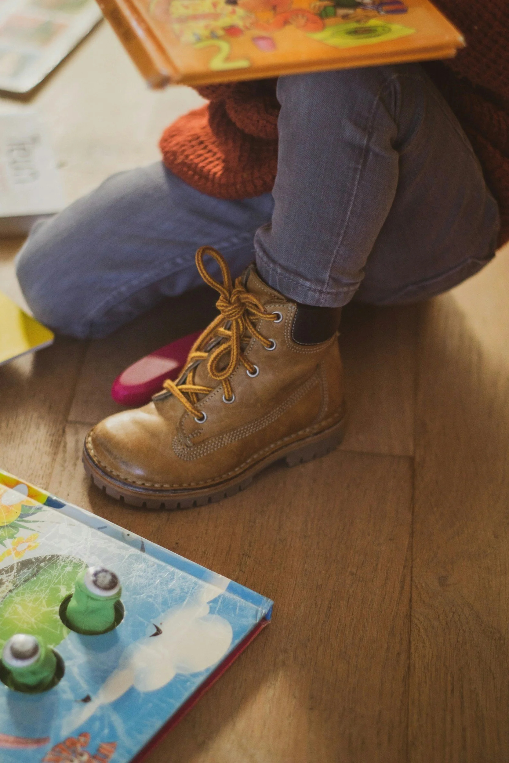 Close-up of a child's leg wearing a brown leather boot, jeans, and a rust-colored sweater, sitting on a wooden floor next to a colorful board game.