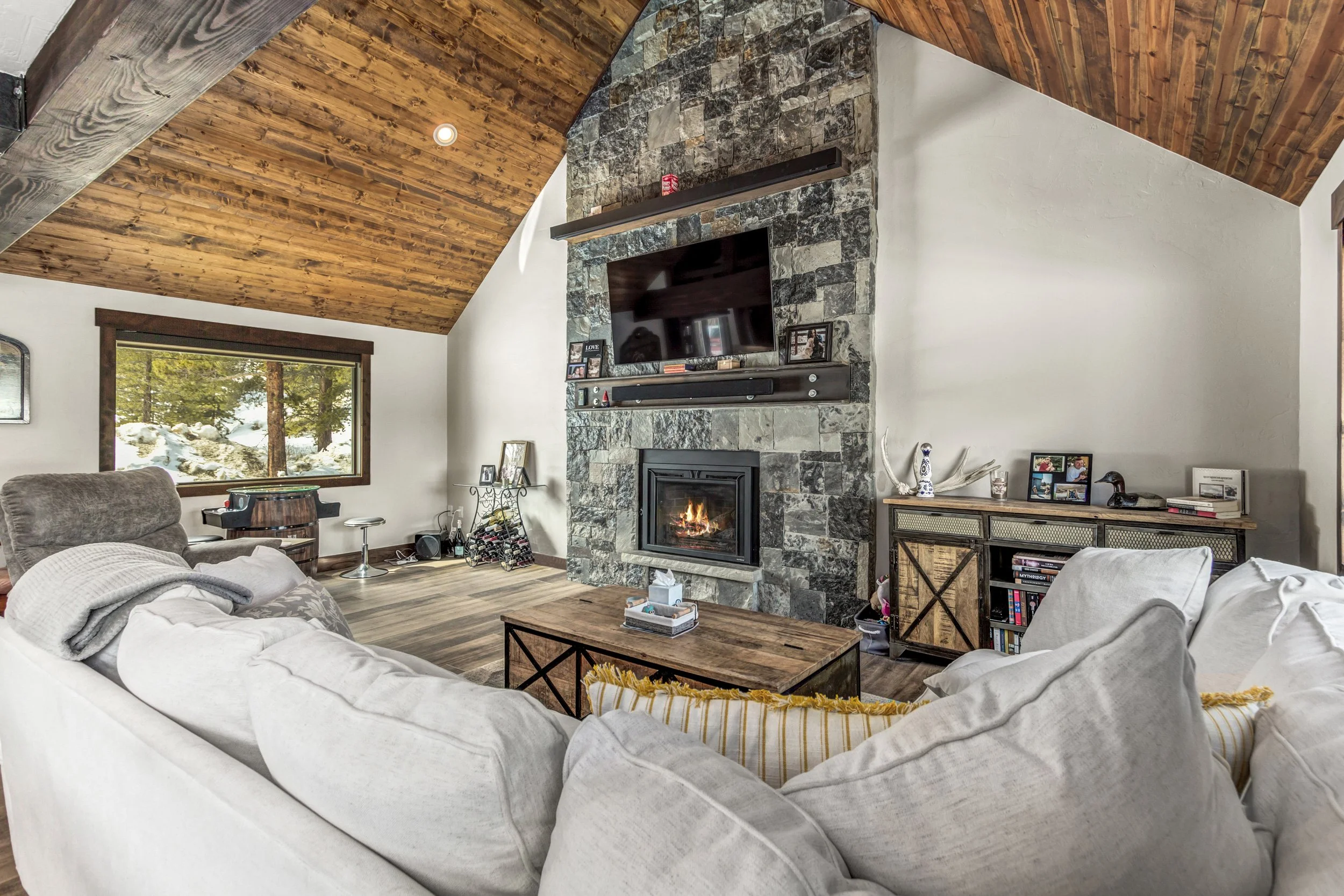 Living room with a white sectional sofa, a stone fireplace with a mounted TV above, a large window showing trees and snow outside, and a wooden ceiling with exposed beams.