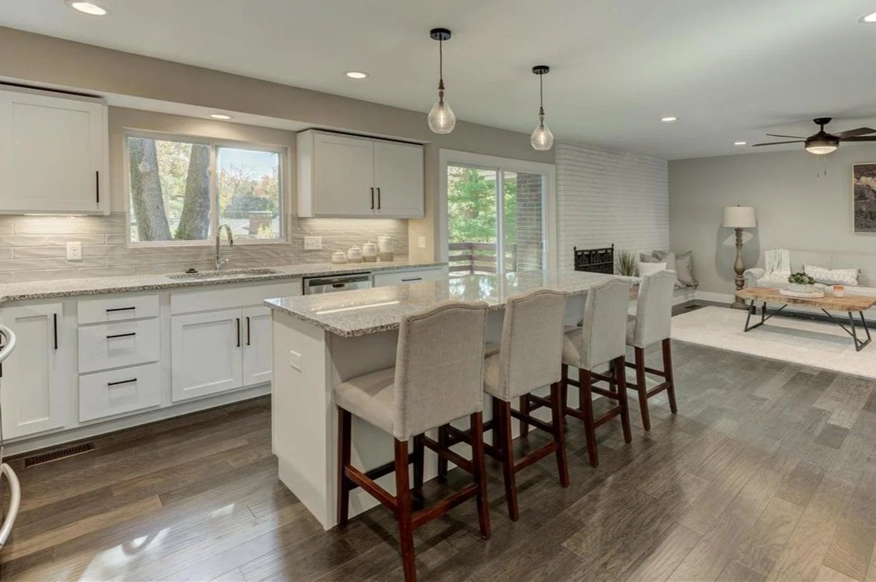 Open-concept kitchen and living room with white cabinets, granite countertops, a kitchen island with four beige upholstered chairs, hardwood flooring, a sliding glass door, a ceiling fan, a sofa with pillows, a coffee table, and a floor lamp.