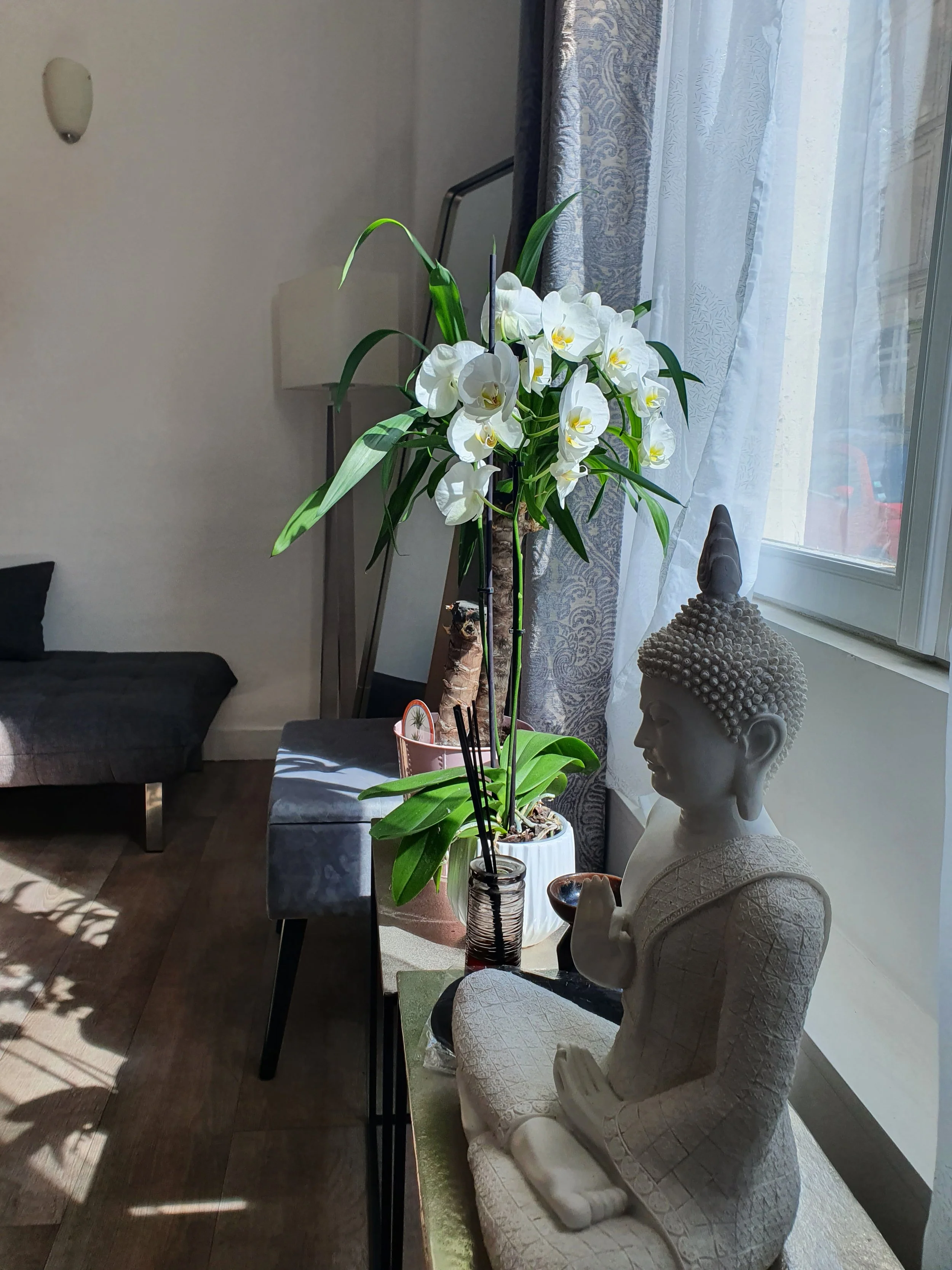 Statue de Bouddha en pierre blanche dans un intérieur, avec un vase de fleurs blanches sur la table à côté, près d'une fenêtre avec rideaux.
