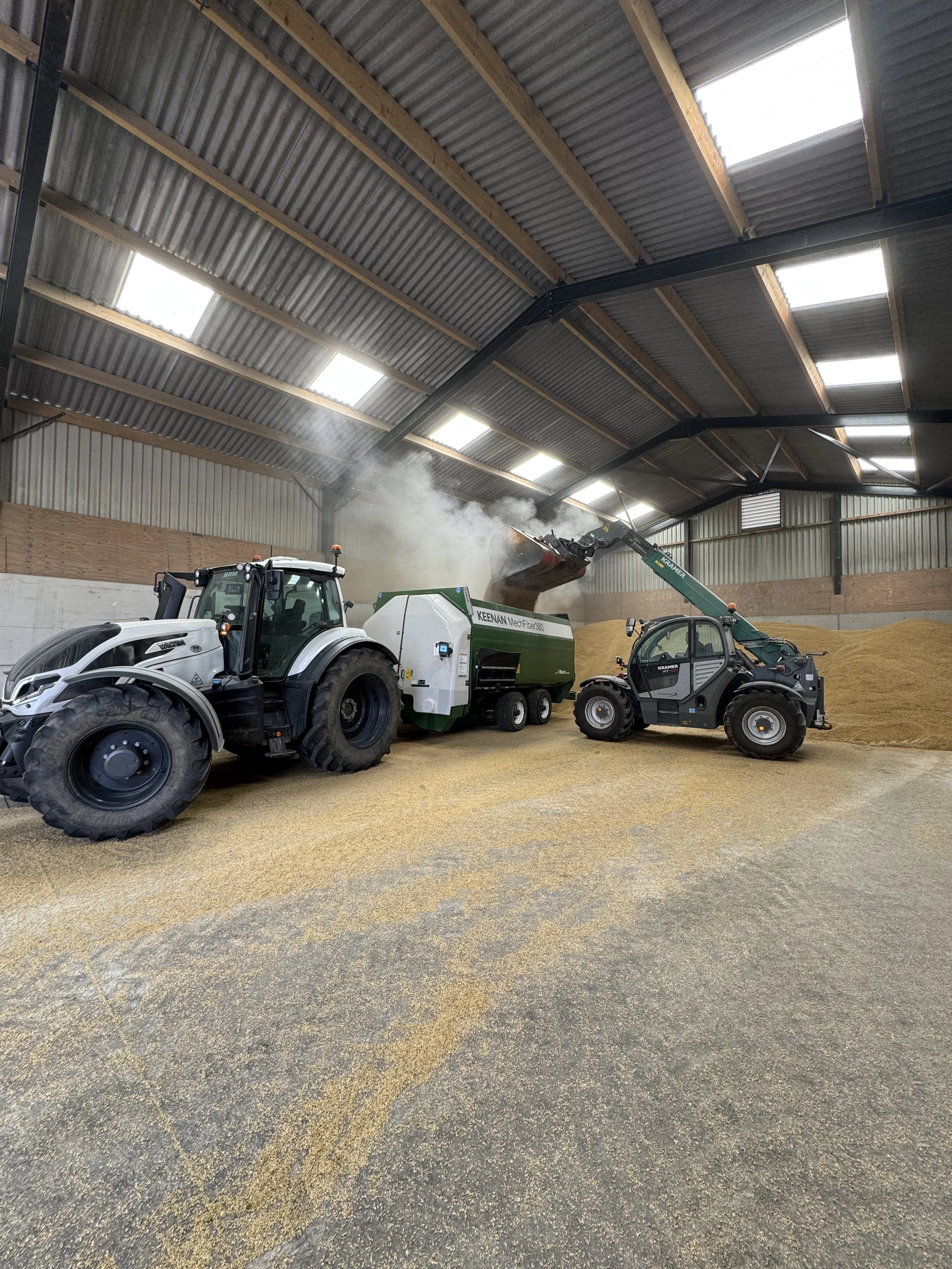 Inside an industrial warehouse with a metal roof, a tractor and a smaller utility vehicle are parked on a concrete and gravel floor, with a large green and white machine pouring a substance into a pile of grain or feed.
