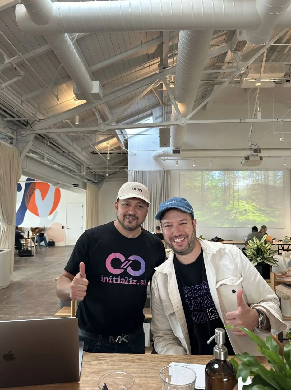 Two men smiling and giving thumbs-up while standing in a modern, well-lit indoor space with a high ceiling, large windows, and a projector screen displaying a forest scene behind them.