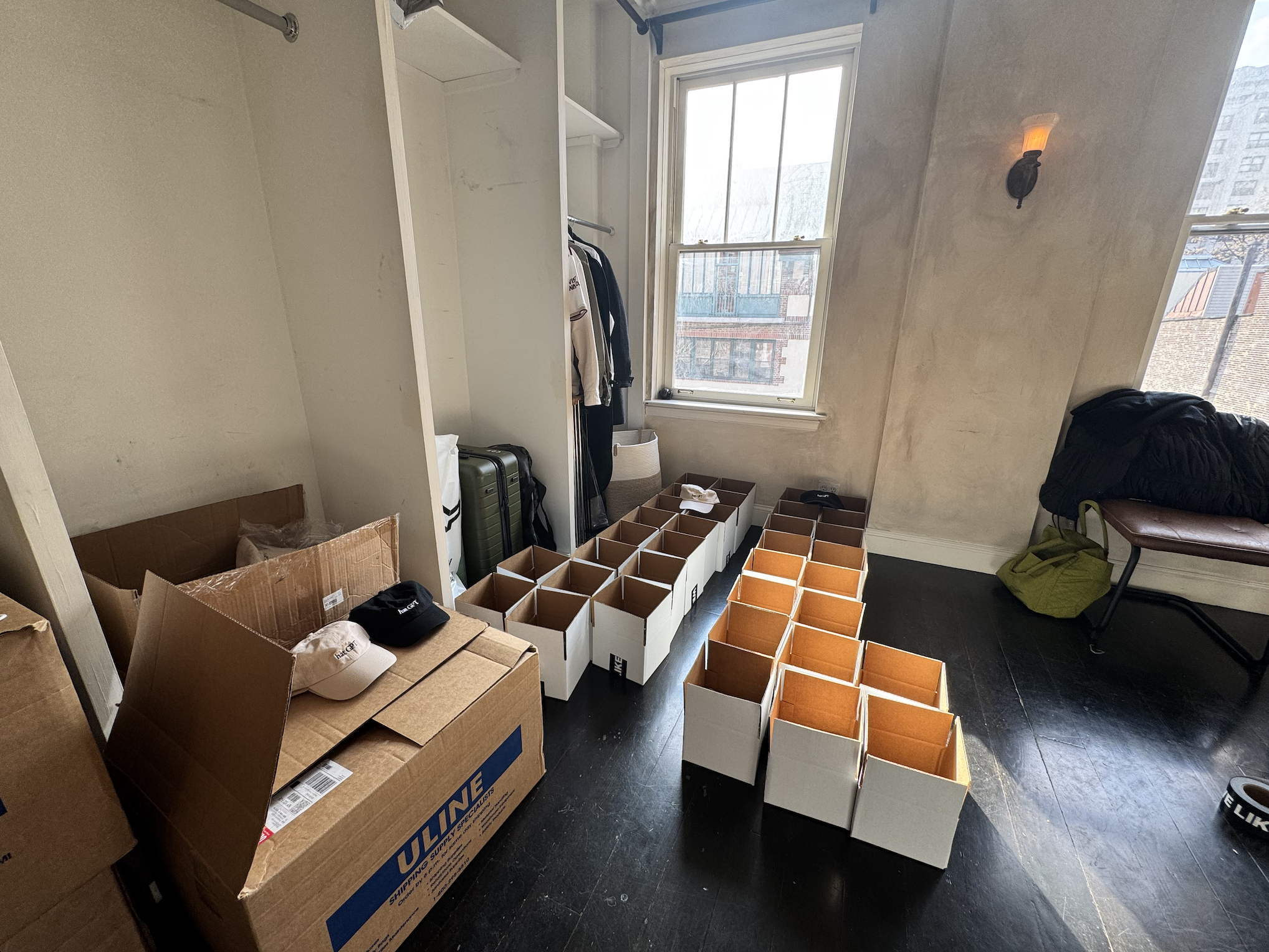 Room with open closet, cardboard boxes, and empty white storage bins with orange interiors on the dark wooden floor. Contains a window, some clothing, a chair, and a black and green bag.