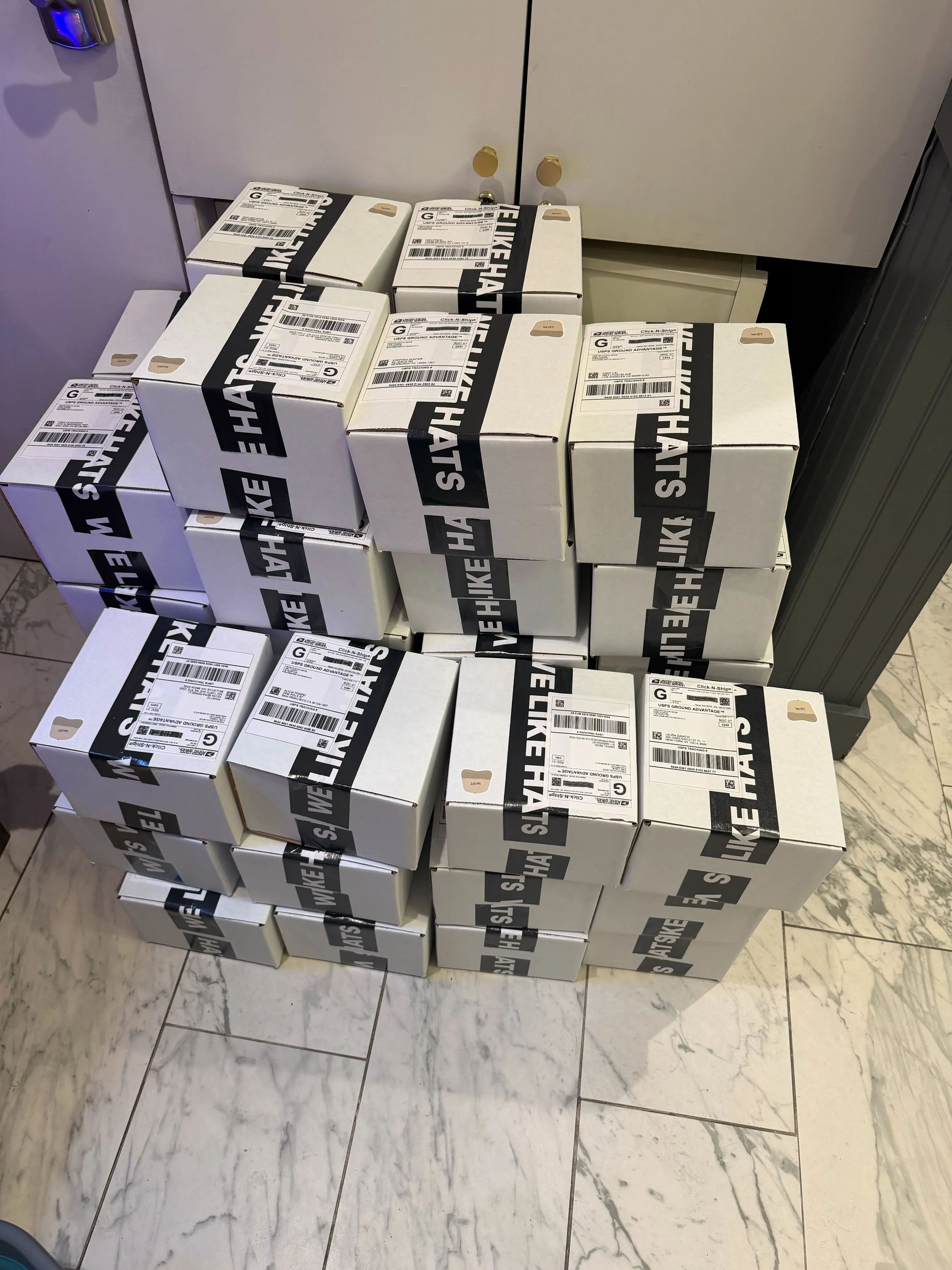 Multiple white boxes with black tape that reads 'WE LIKE HATS' stacked on a marble floor near a stainless steel door.