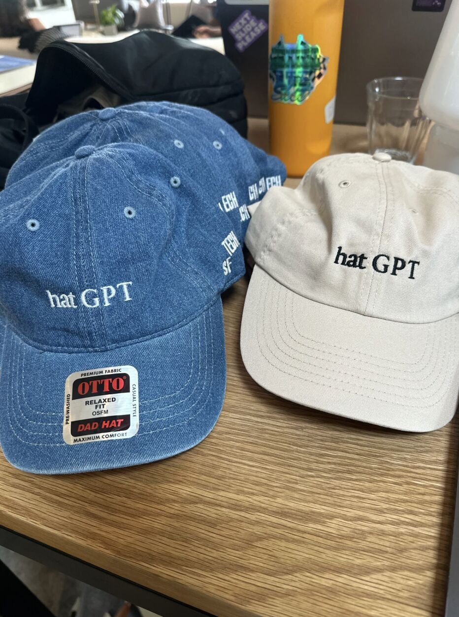 Two baseball caps, one blue and one beige, both embroidered with 'hat GPT,' placed on a wooden table. The blue cap has a sticker that reads 'OTTO relaxed fit dad hat.' In the background, there are various objects including a yellow beverage bottle, a clear glass, and some unidentified items.