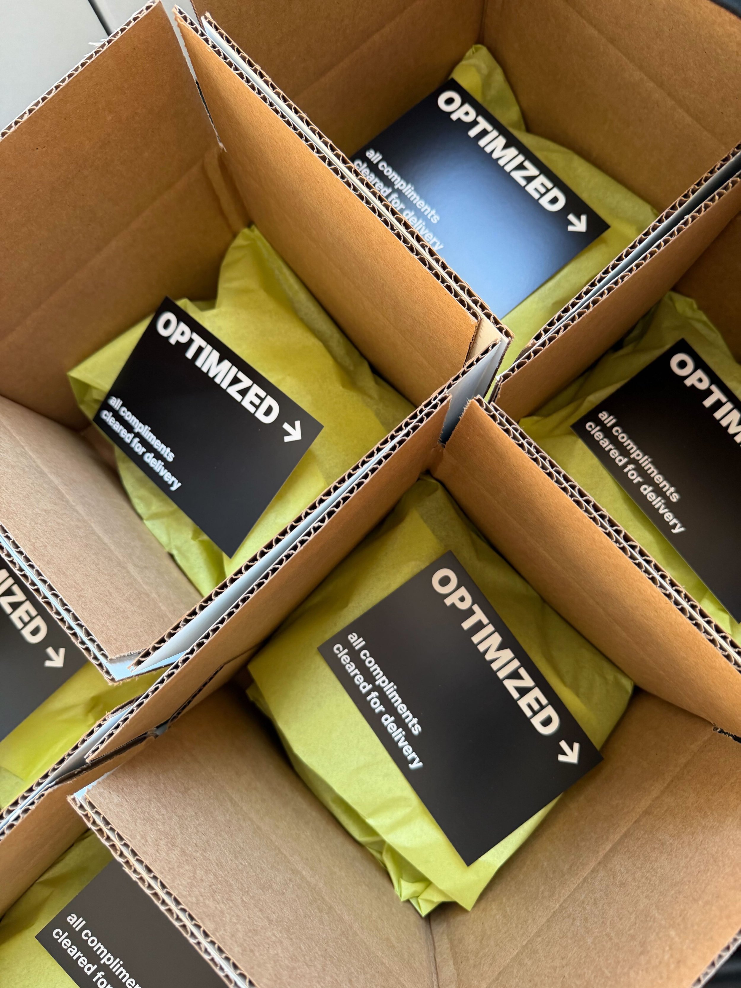 Inside a cardboard box with four compartments, each containing yellow tissue paper and a black card labeled 'OPTIMIZED' with an arrow and the text 'all compliments cleared for delivery'.