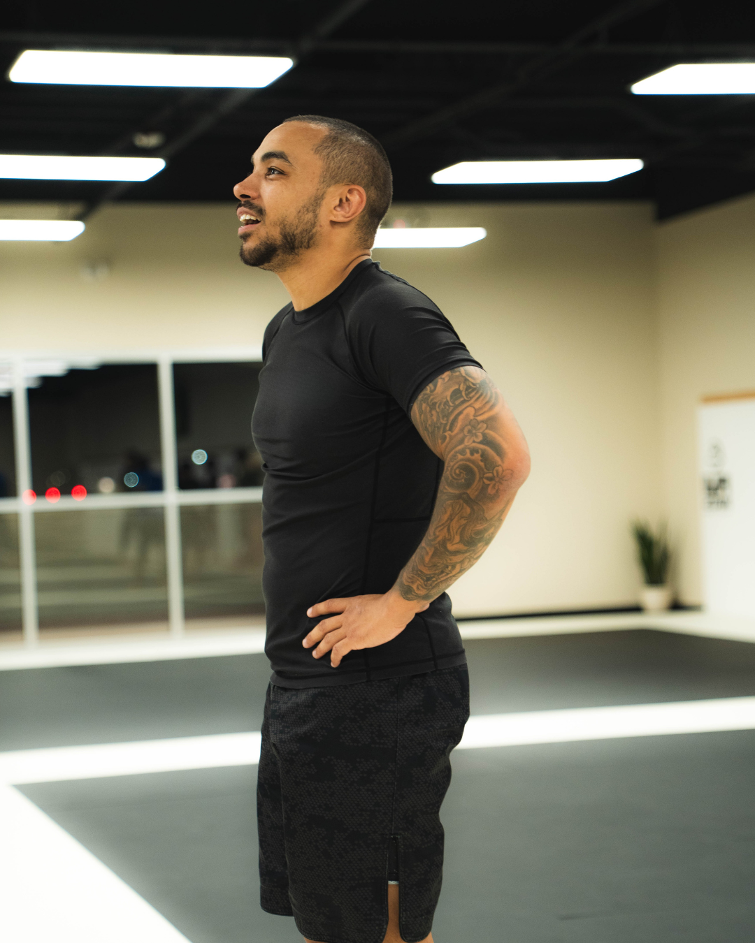 A man with a tattooed arm and a beard stands in an indoor gym, wearing a black athletic shirt and shorts, smiling while looking to his left.