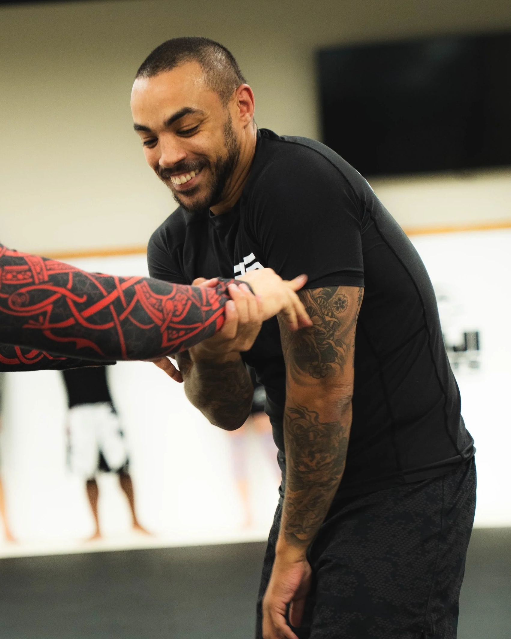 A man with tattoos on his arm smiling and engaging in martial arts training, holding an opponent's arm, in a gym or martial arts studio.