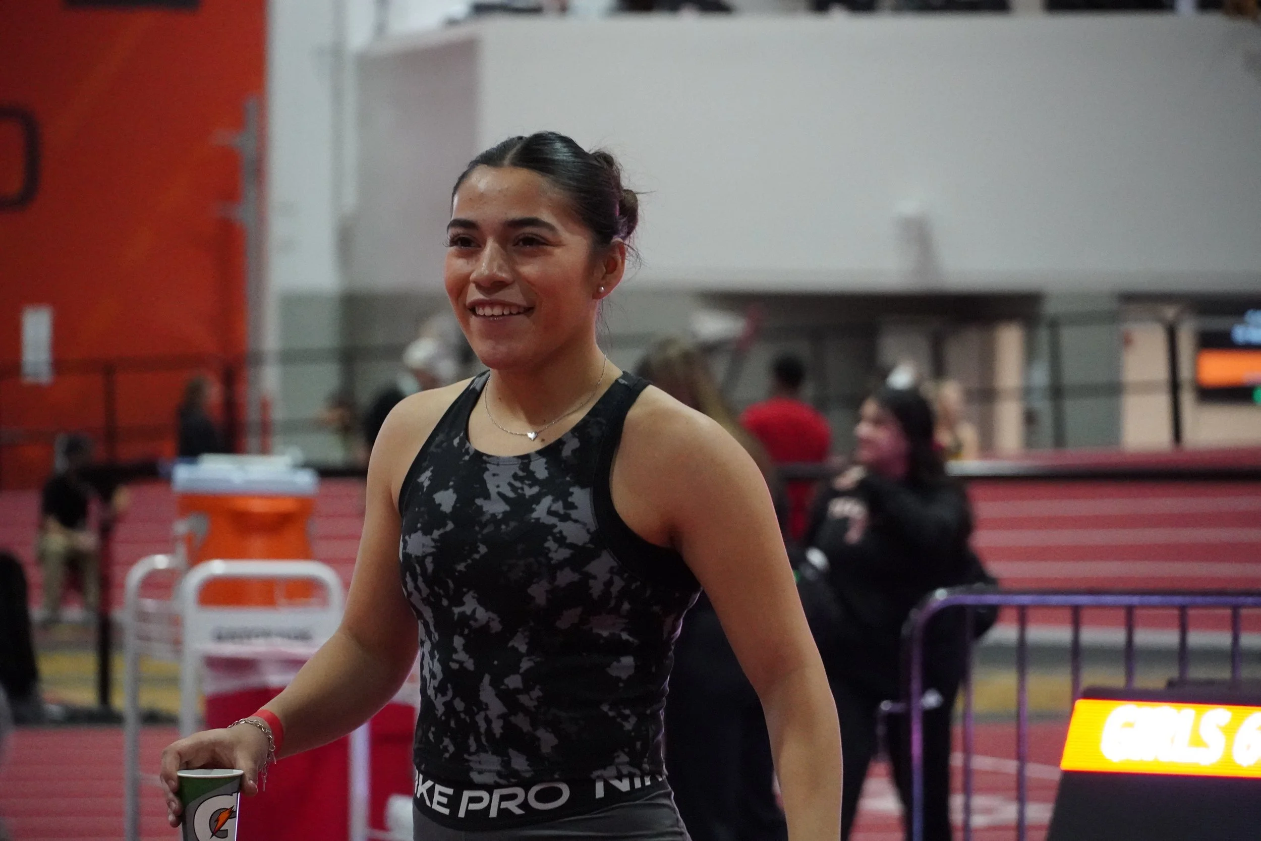 Young female athlete with dark hair in a bun, wearing a black and gray sports tank top, holding a Gatorade cup, smiling at an indoor track and field event.