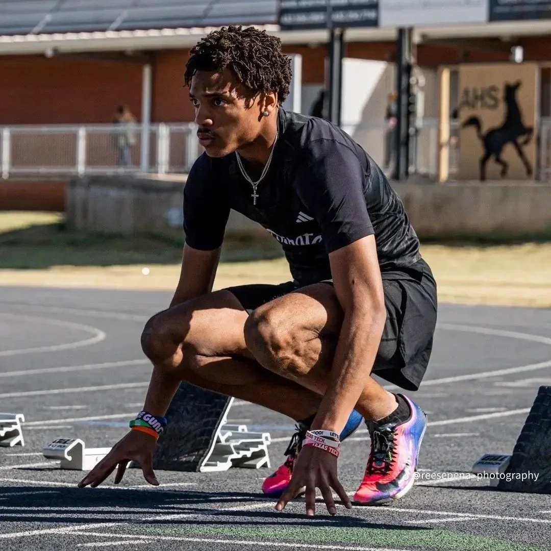 A young male athlete crouching at a starting line on an outdoor track, preparing for a race, wearing athletic clothing and colorful running shoes.