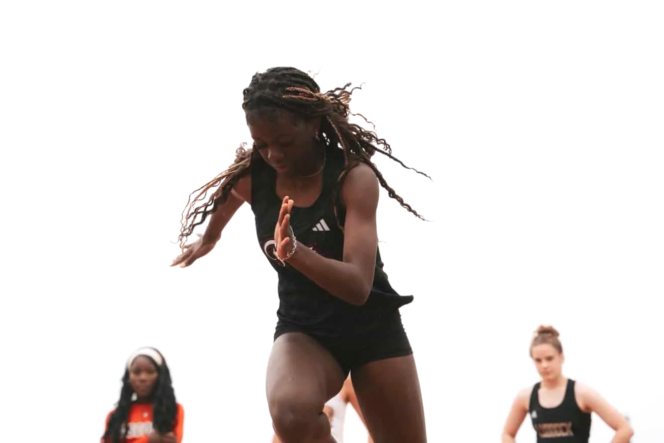 A woman with braided hair, wearing a black athletic tank top and shorts, is jumping or running against a white background, with two women in similar athletic outfits in the background.