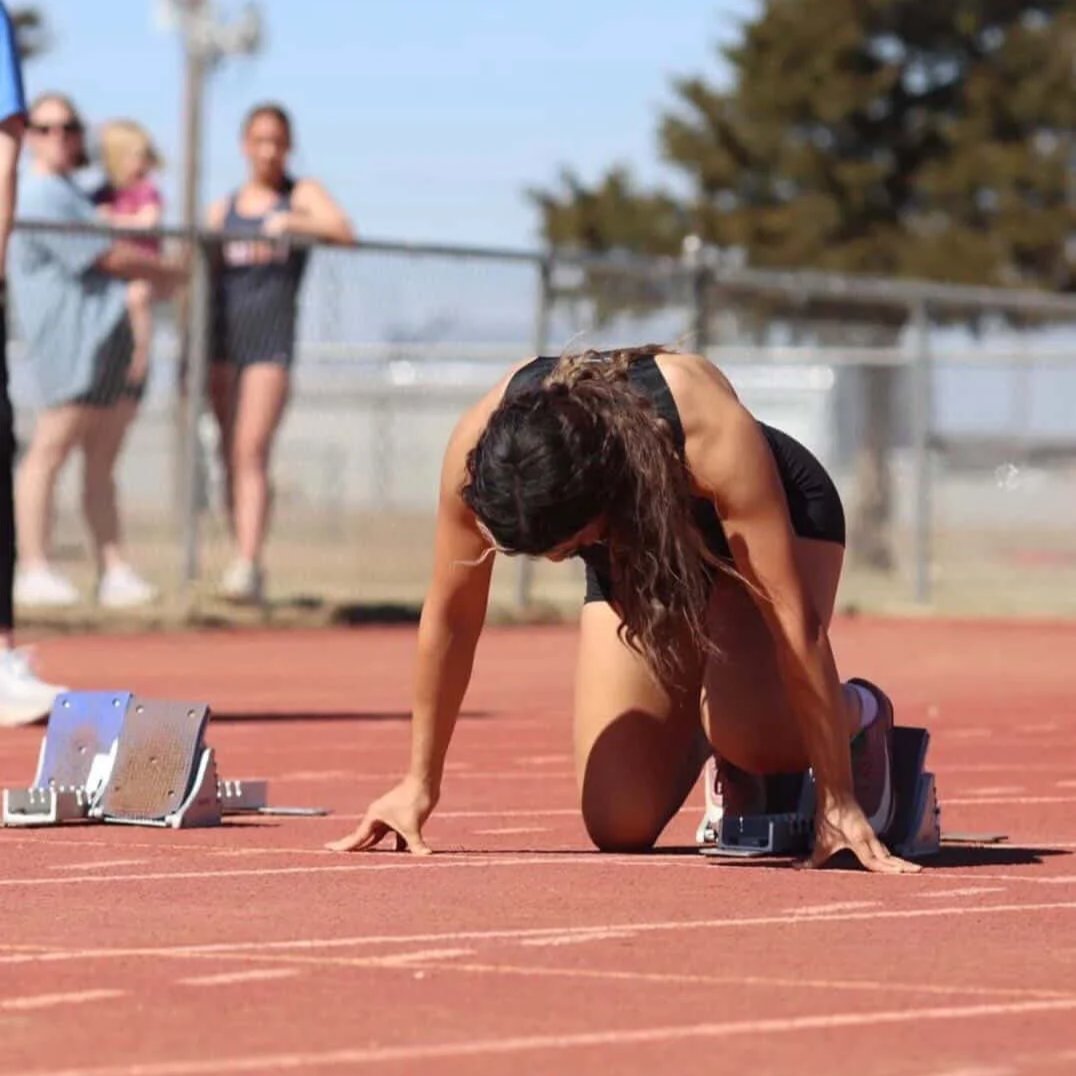 Female athlete in starting position on a running track at a track and field event, with a few spectators in the background.