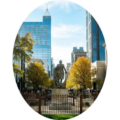 A bronze statue of a man in a city park, surrounded by trees with yellow leaves, with tall modern skyscrapers in the background.