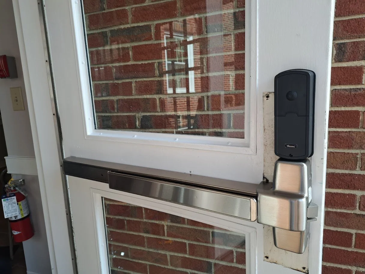 Exterior door with a black electronic lock and a stainless steel push bar, set against a brick wall with a window.
