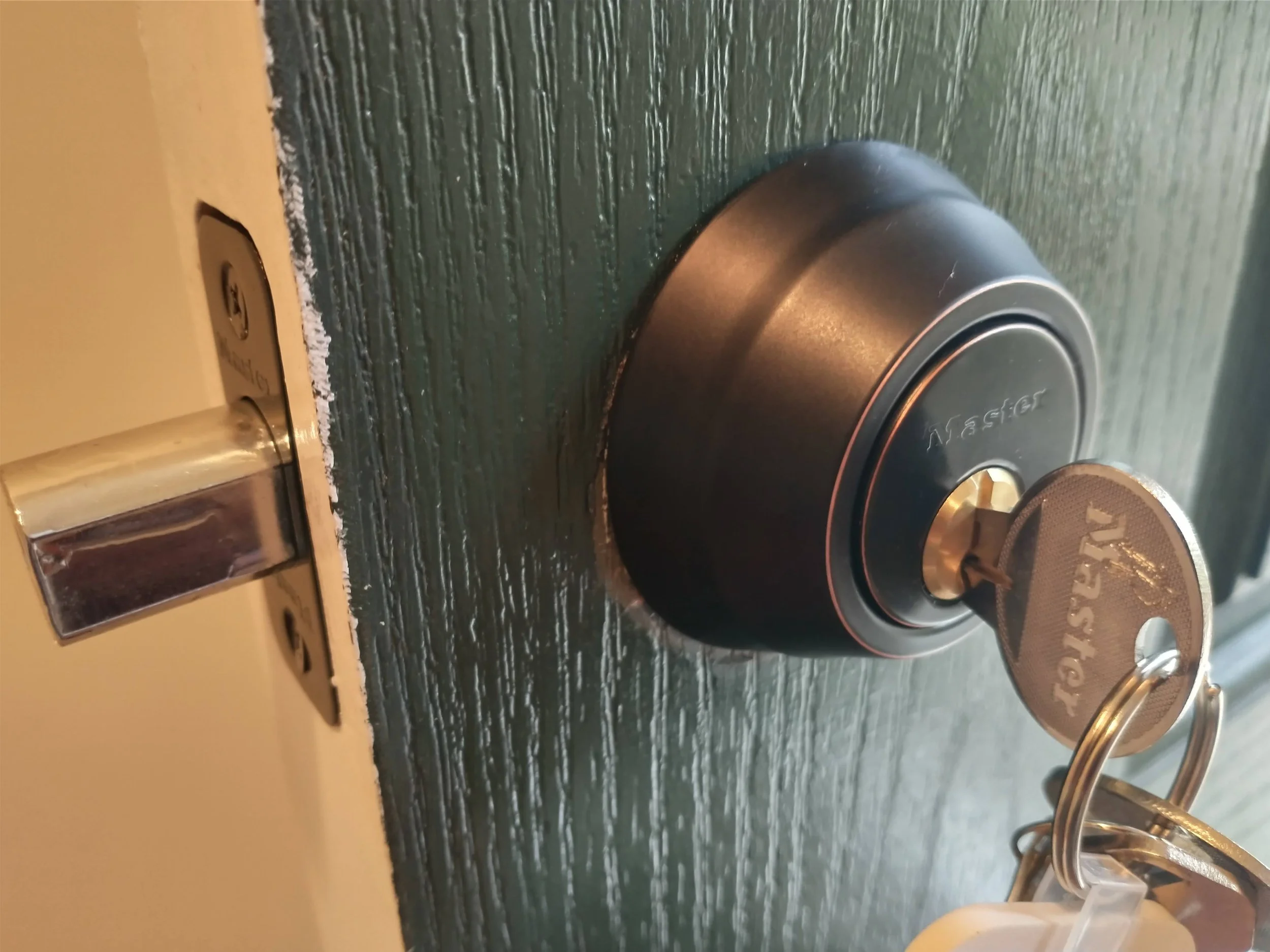 A black Master Lock padlock with a key inserted and a keychain attached, placed on a dark green textured wood surface. The keychain has a circular metal tag with the brand name 'Master' engraved on it.