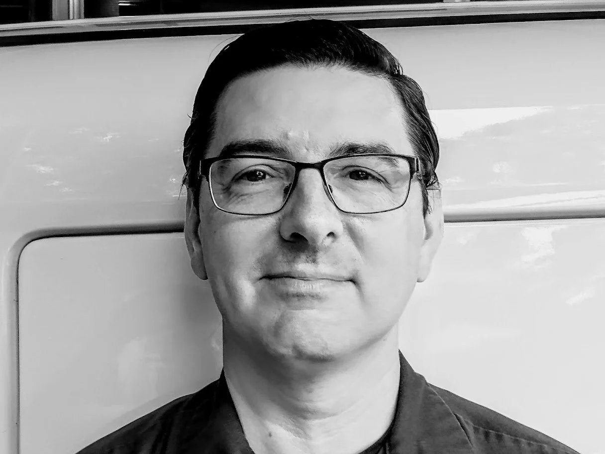 A black and white photo of a middle-aged Asian man with short dark hair, wearing glasses and a collared shirt, standing in front of a light-colored wall.