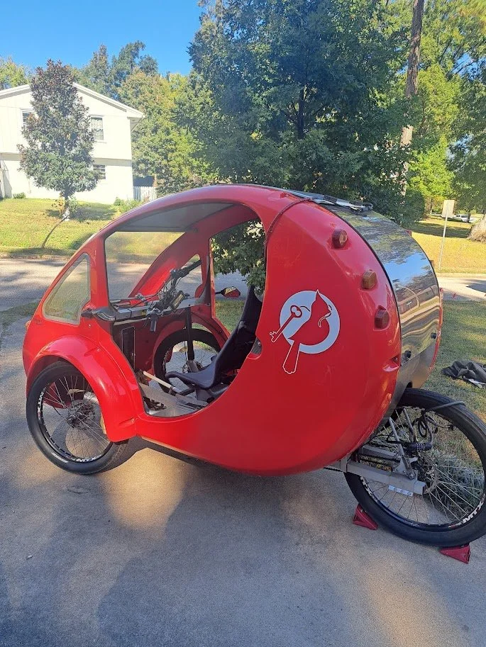 A red velomobile with a shell that has a logo of a dog with a wrench and a screwdriver, parked on a driveway with trees and a house in the background.