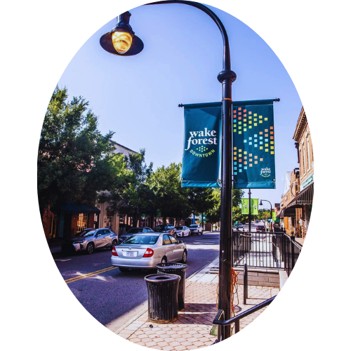 Street scene with a street lamp and banner that says 'Wake Forest Downtown.' There are cars parked along the street and buildings on both sides, with trees lining the sidewalk.