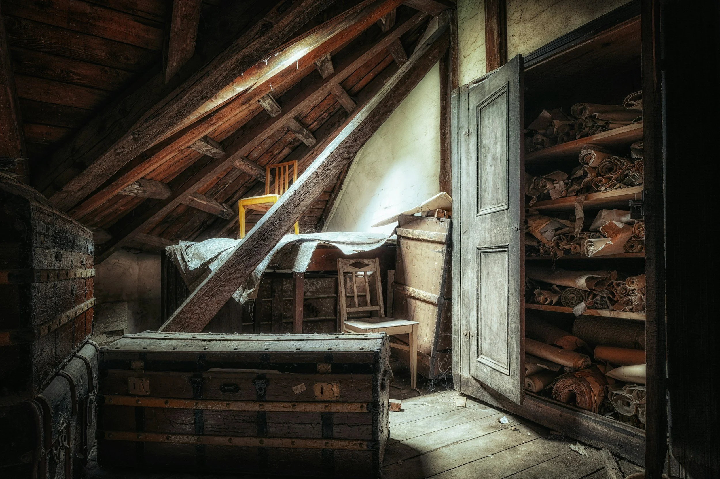 An attic room filled with old furniture, including a yellow chair on a sloped ceiling, a wooden chest, a small table with a chair, and a large open cabinet filled with rolled-up papers. The room appears dusty and abandoned with cobwebs and debris.