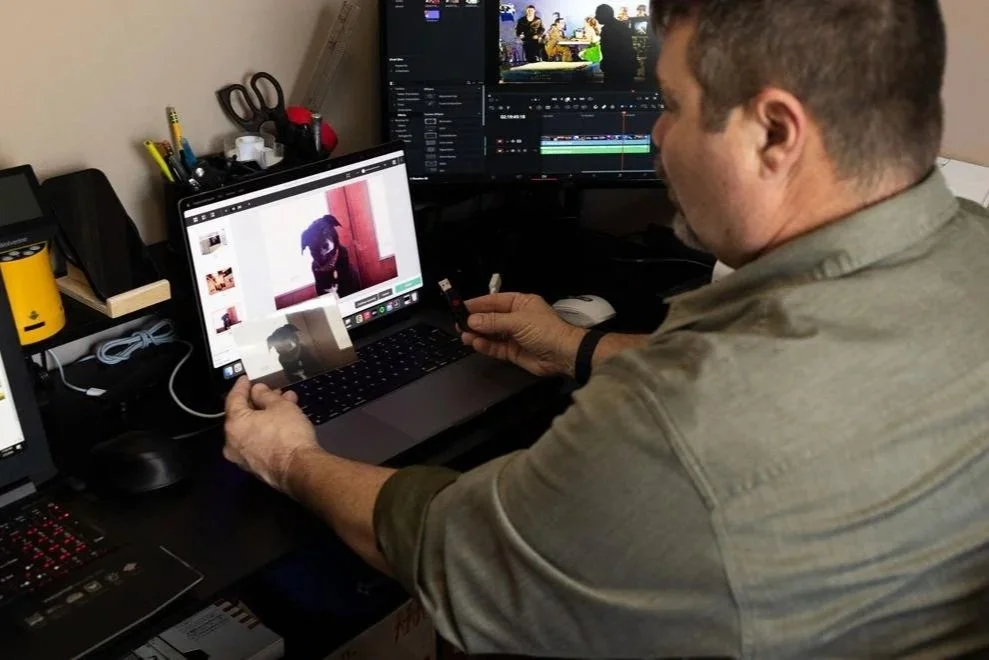 A man sitting at a desk editing a video on a large monitor and holding a printed photo of a dog sitting on a windowsill. The desk has a laptop, scissors, pens, and other supplies.