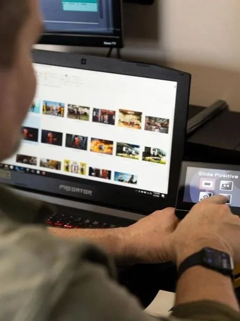 Person using a smartphone to select a positive slide for a presentation in front of a computer monitor showing photos or videos.