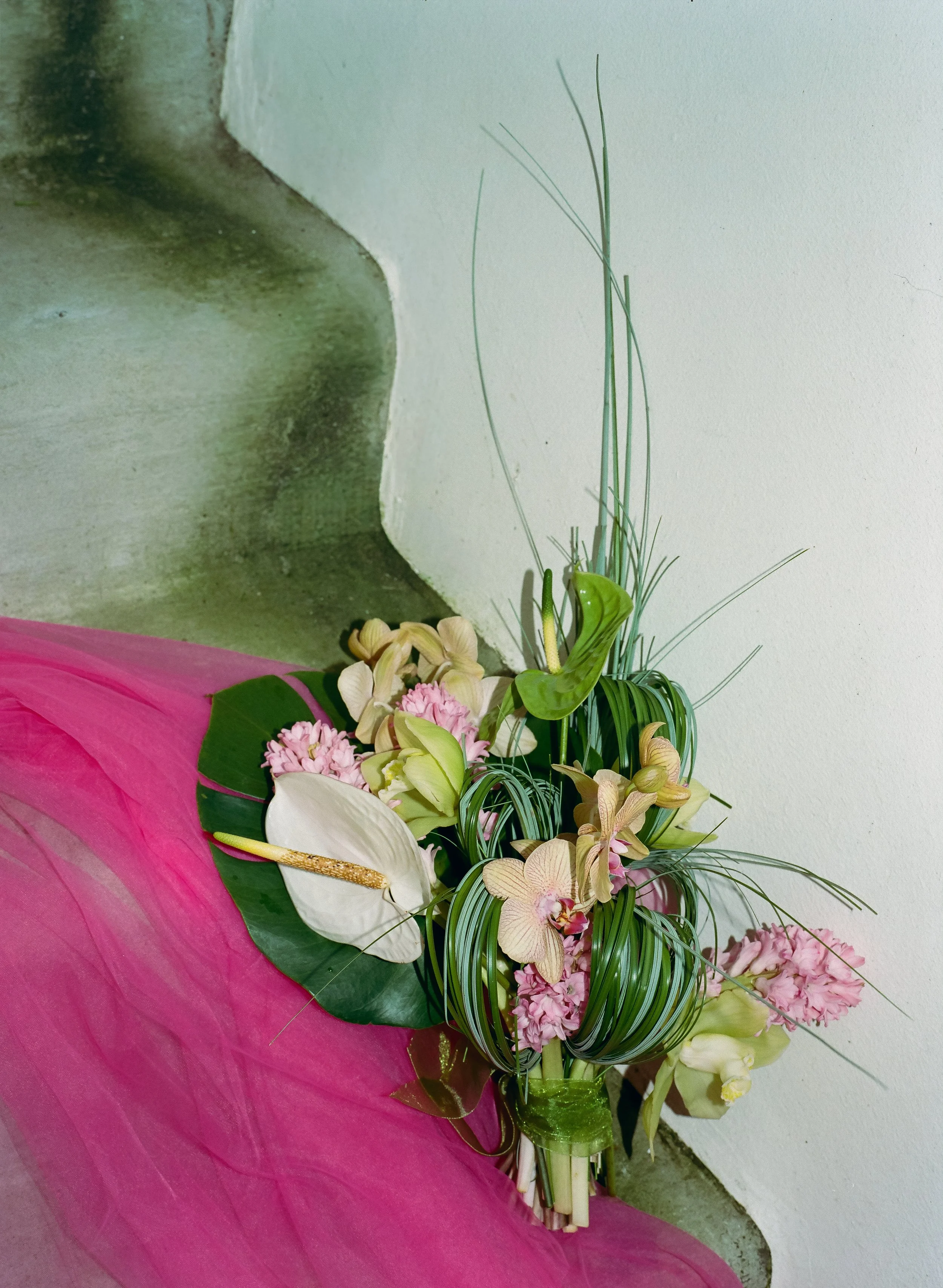 A bouquet of mixed flowers including white calla lilies, pink and cream carnations, and orchids, wrapped in pink and green paper, leaning against a wall on a concrete floor.