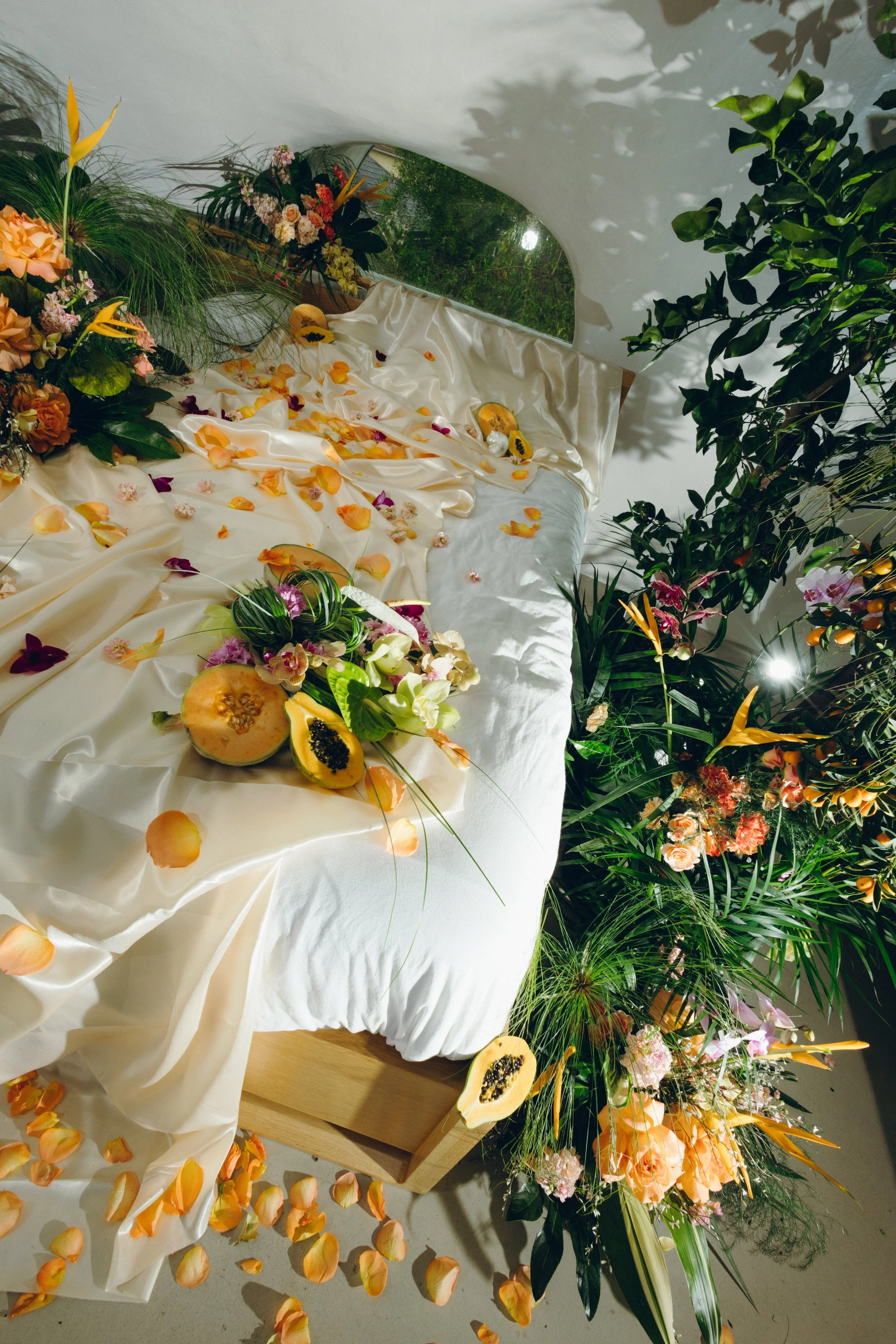 A floral display with various flowers and greenery on a white satin draped surface, with flower petals scattered around, set near a window with a view of trees outside.