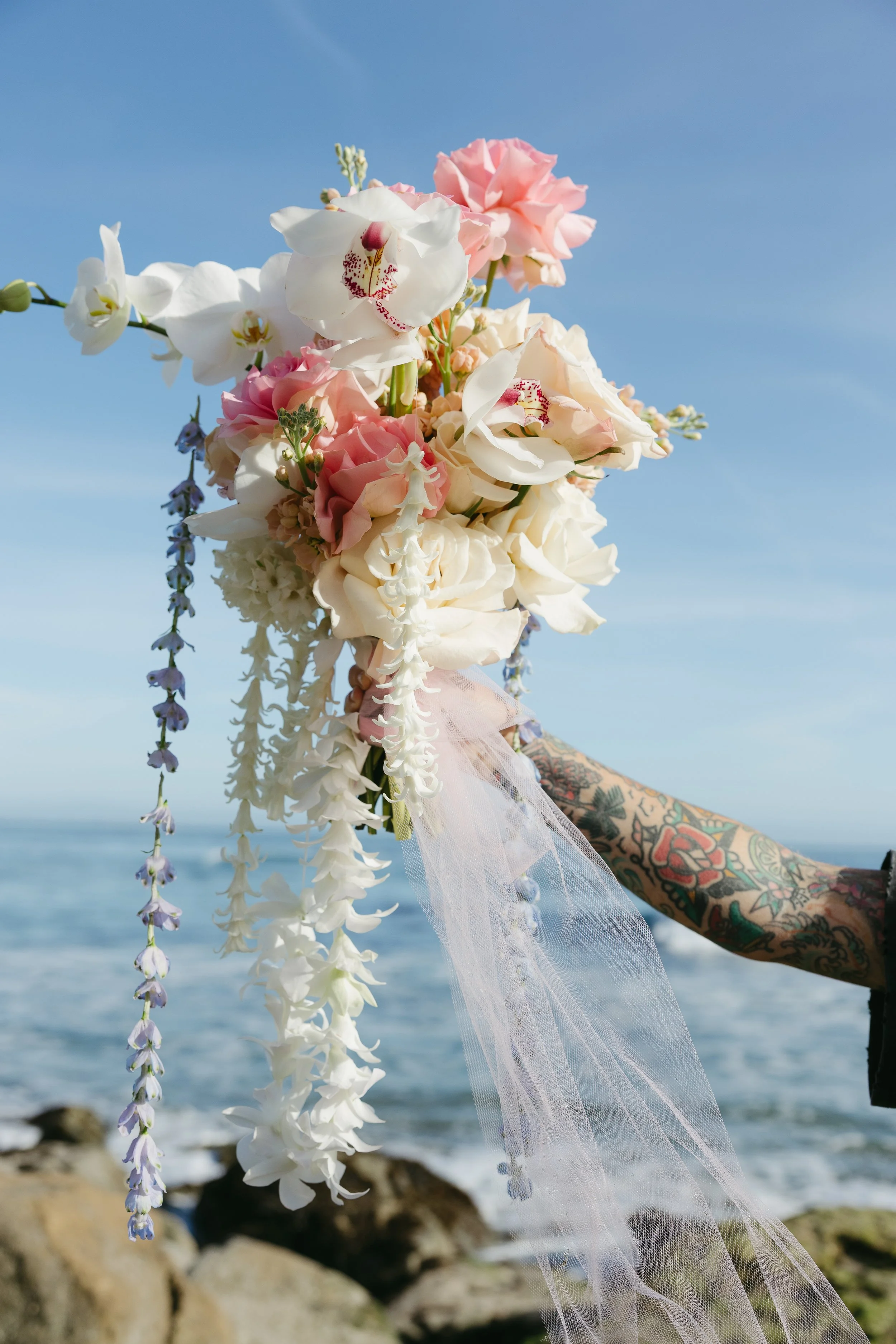 Bouquet of pink and white flowers being held by tattooed hand with a blue sky and ocean in the background.