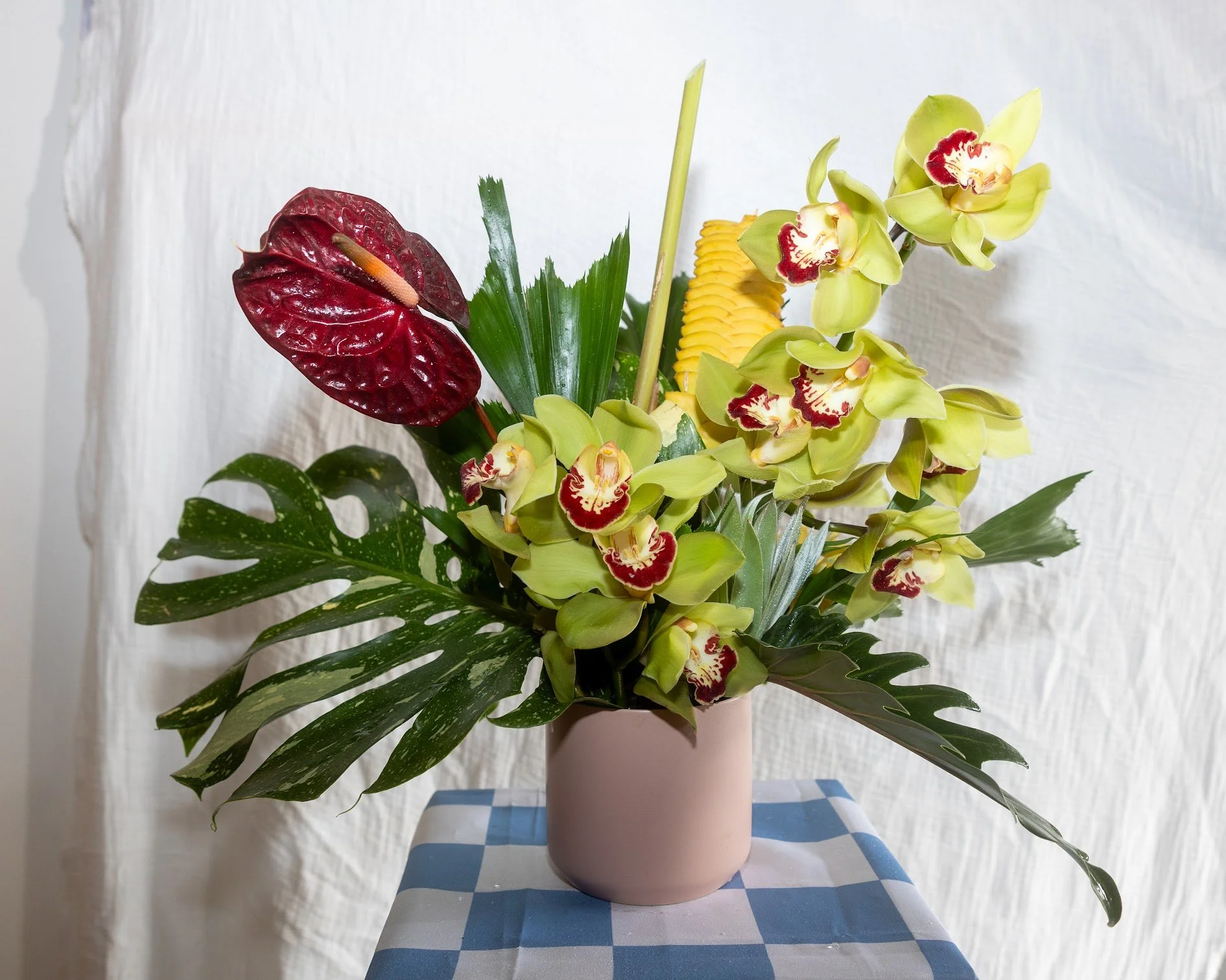 A pink vase with a floral arrangement of large green leaves, yellow orchids with red markings, and an anthurium with dark red, heart-shaped leaves and a yellow spadix, on a blue and white checkered tablecloth against a plain white background.