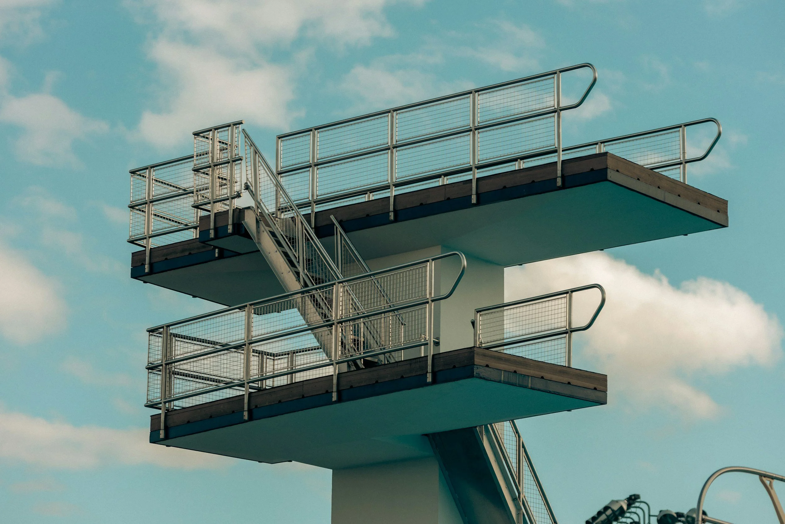 A multi-level outdoor swimming pool diving platform with three sections, each with metal railings, set against a blue sky with scattered clouds.