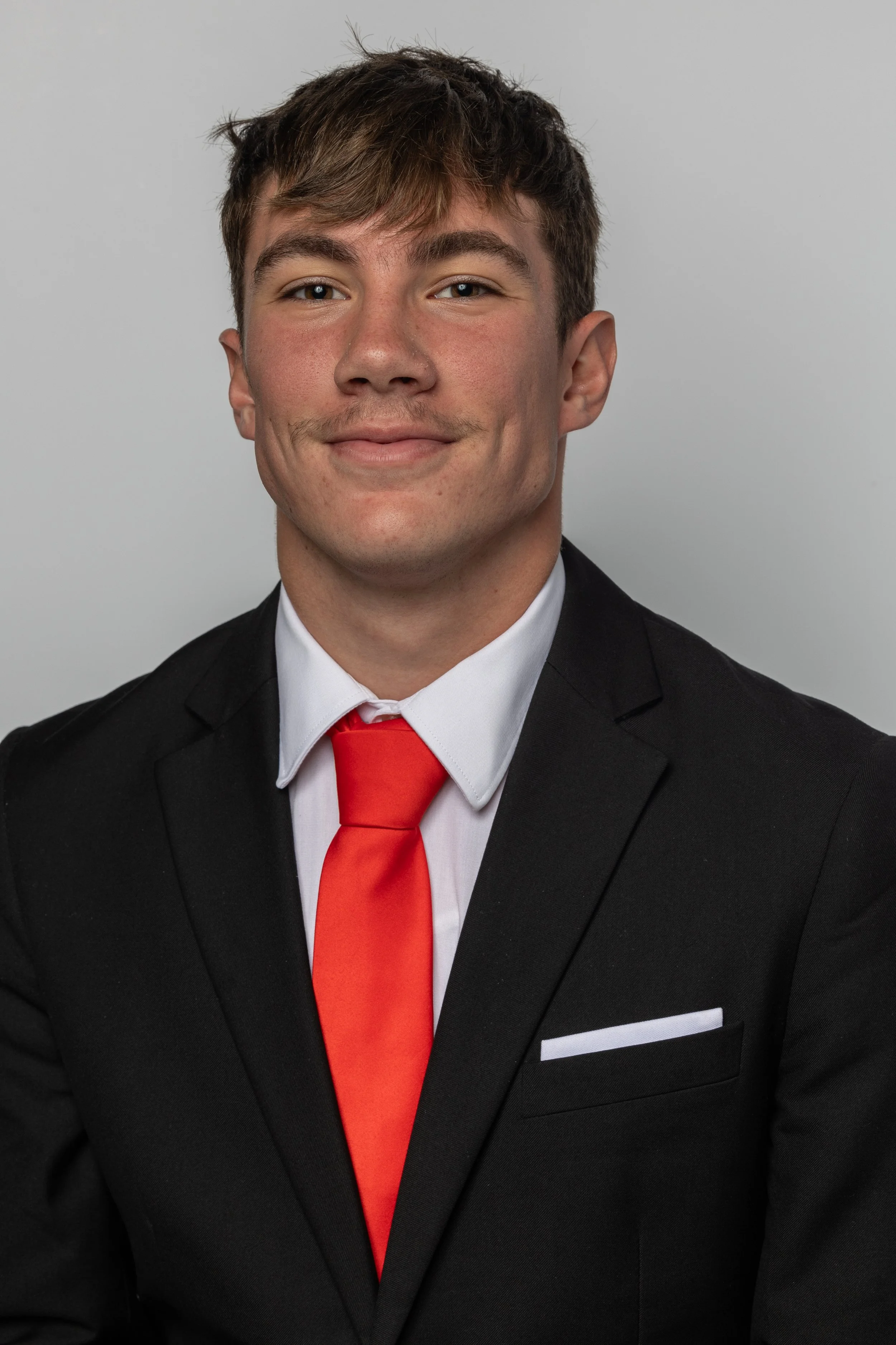 Portrait of a young man in a black suit, white shirt, and red tie, smiling against a plain gray background.