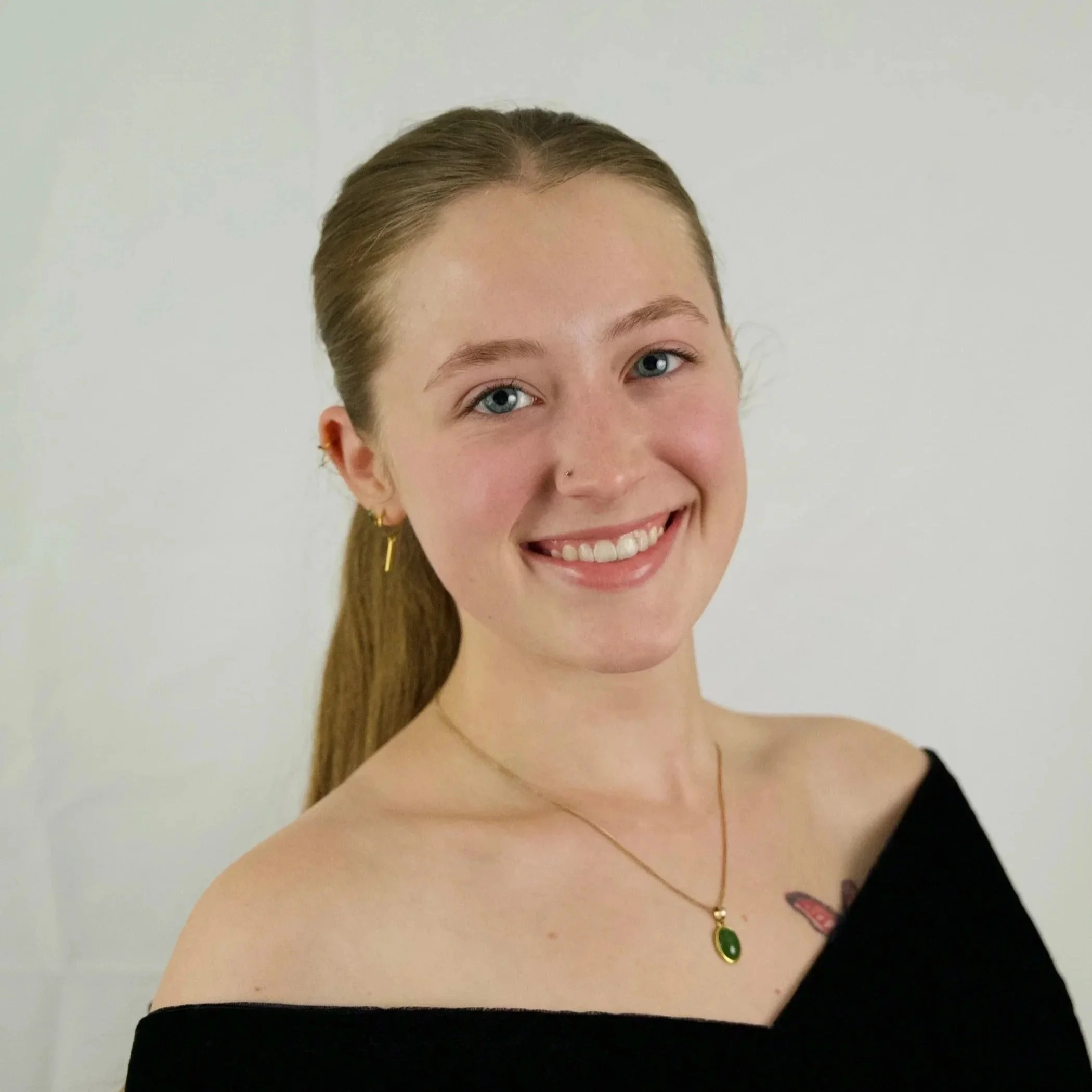 Young woman with light brown hair tied back, wearing a black off-shoulder top, gold jewelry, and a colorful tattoo on her chest, smiling against a plain white background.