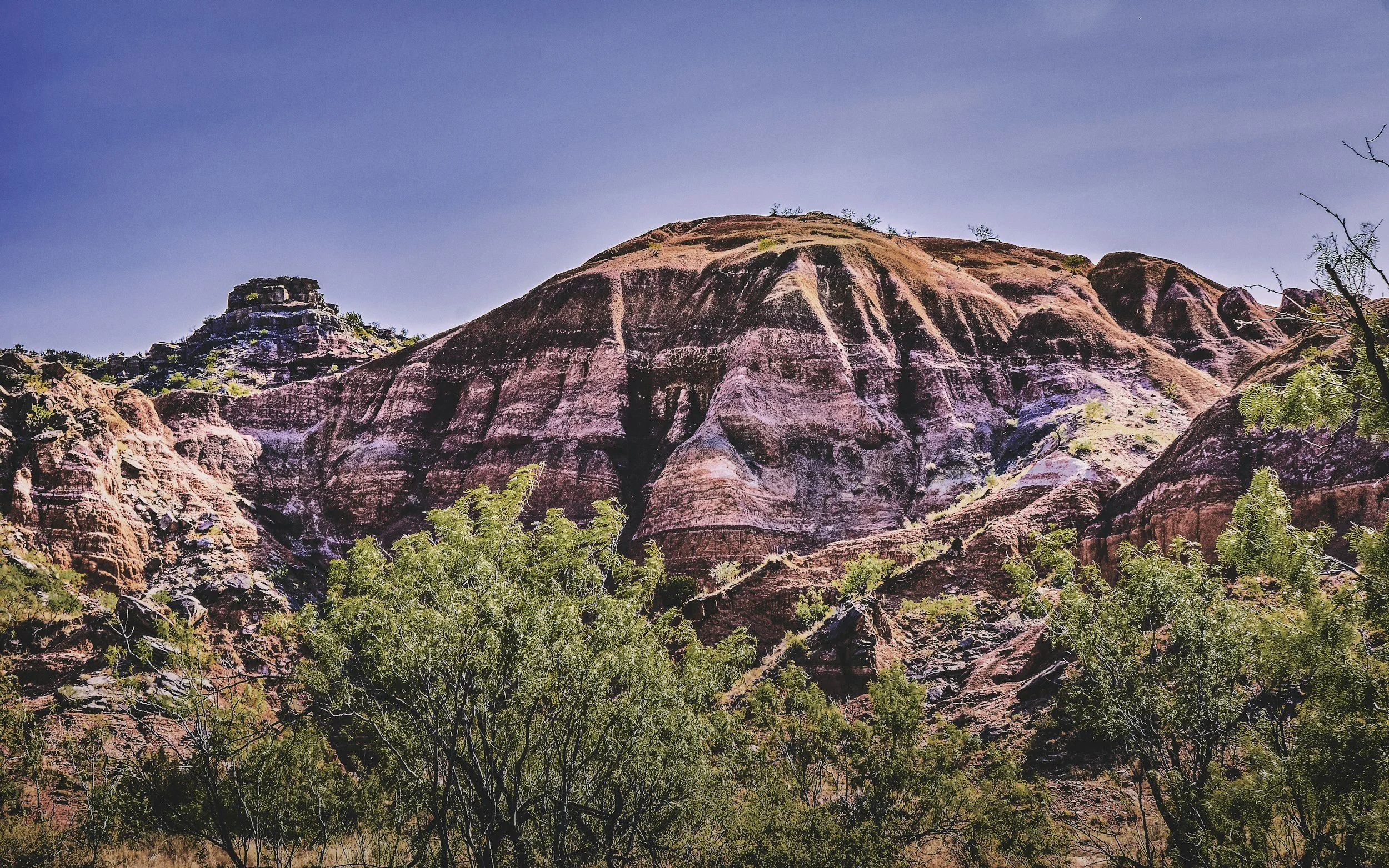 Pink and purple layered rock formations with sparse green vegetation and a clear blue sky.