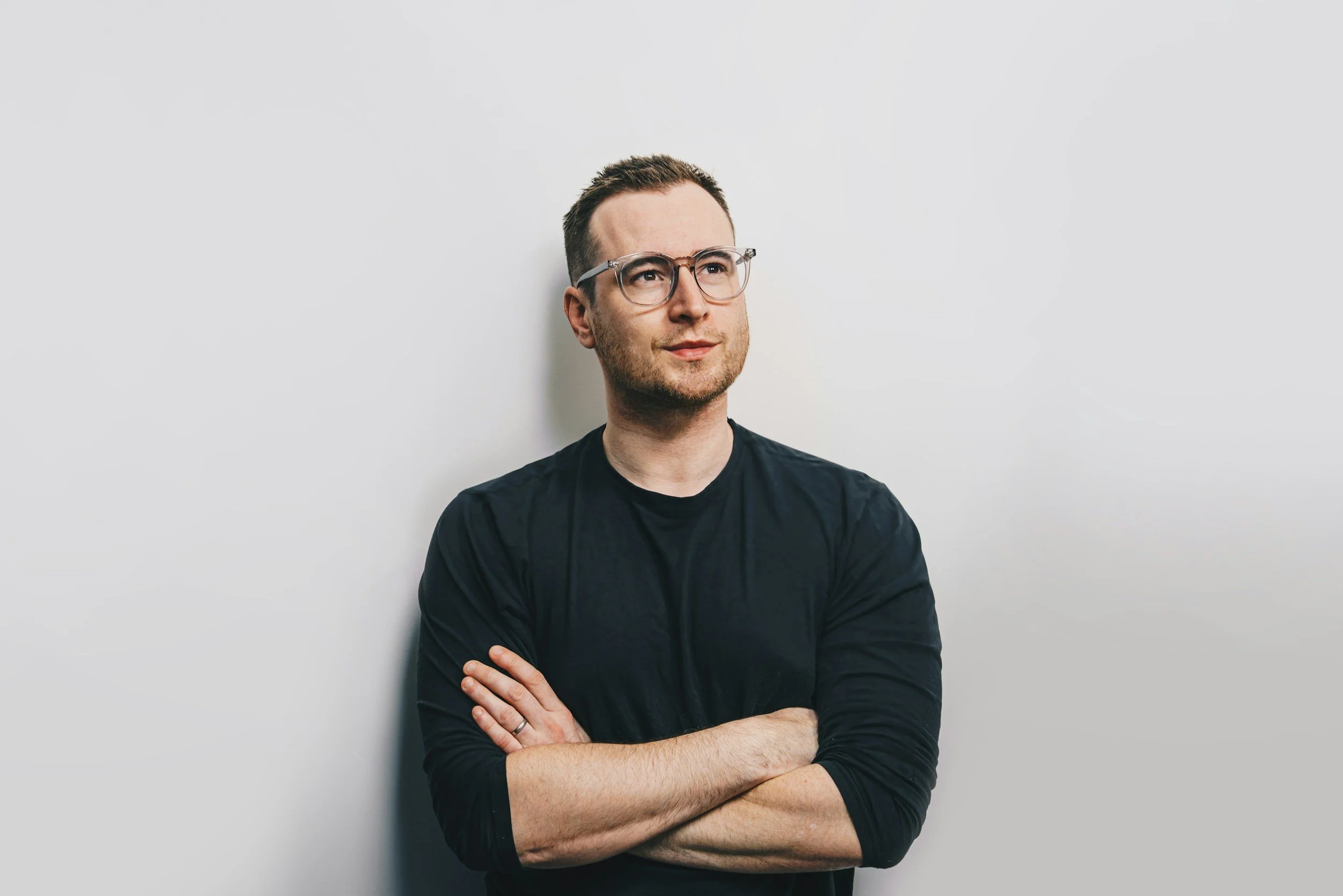 A man with glasses and a black shirt standing against a plain white wall, arms crossed, looking thoughtfully to the side.