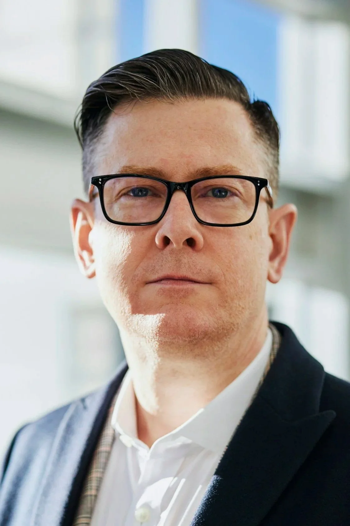 Close-up of a man wearing glasses, a white shirt, and a dark blazer, standing indoors with natural light coming through windows.