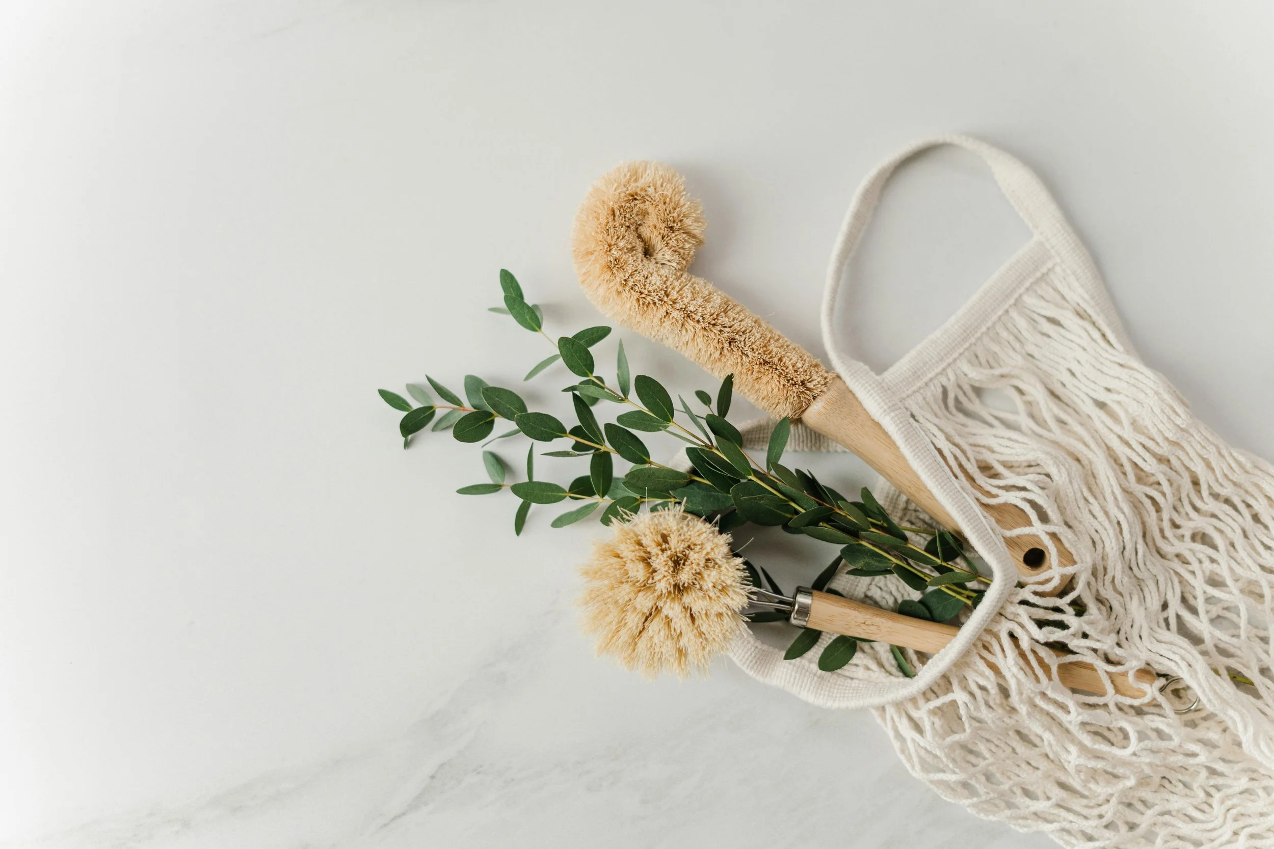 A reusable mesh shopping bag holding two neutral-colored body brushes with wooden handles and a sprig of green leaves, on a white marble surface.