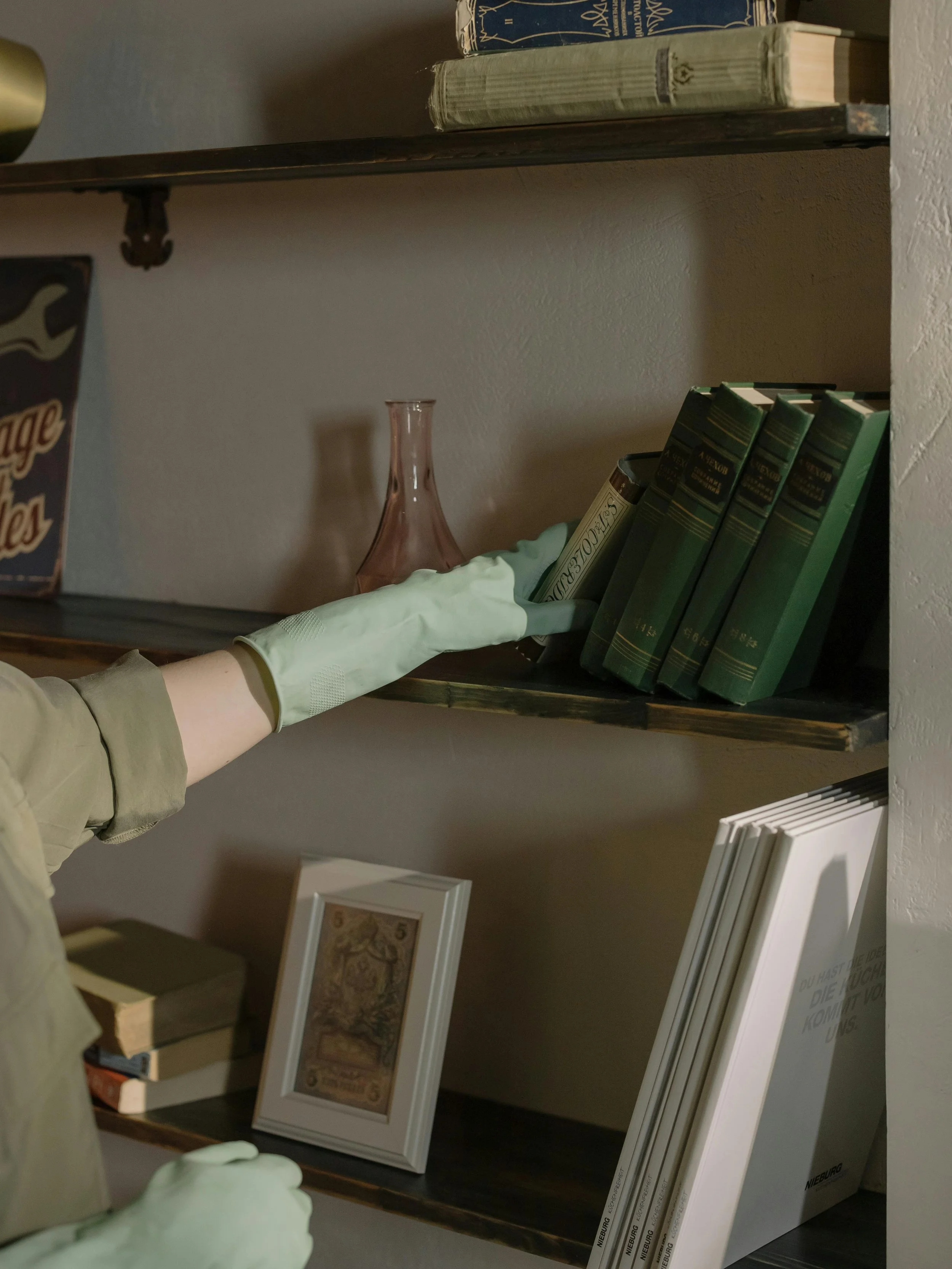 A person wearing a green glove is reaching for a dark green book on a wooden shelf alongside other books and decorative items.