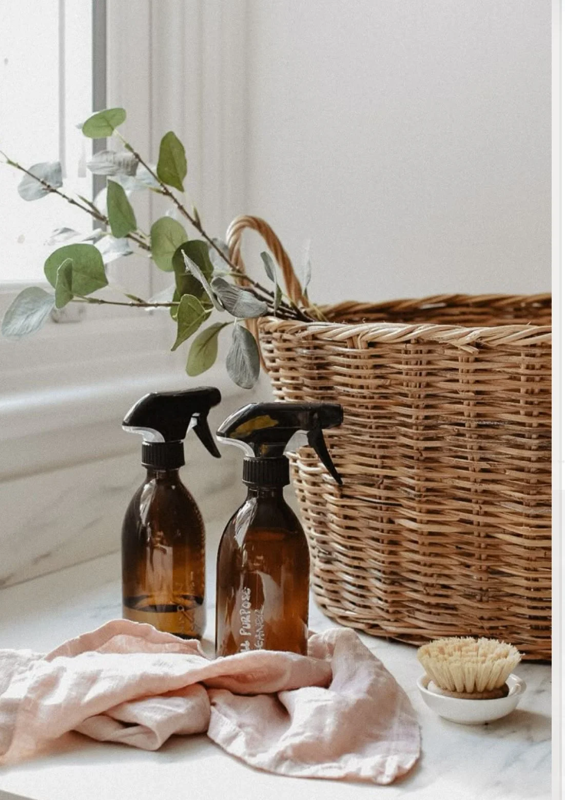 A wicker basket with a green leafy branch inside, two brown spray bottles, a rolled pink cloth, and a small brush in a white dish, all on a white surface near a window.