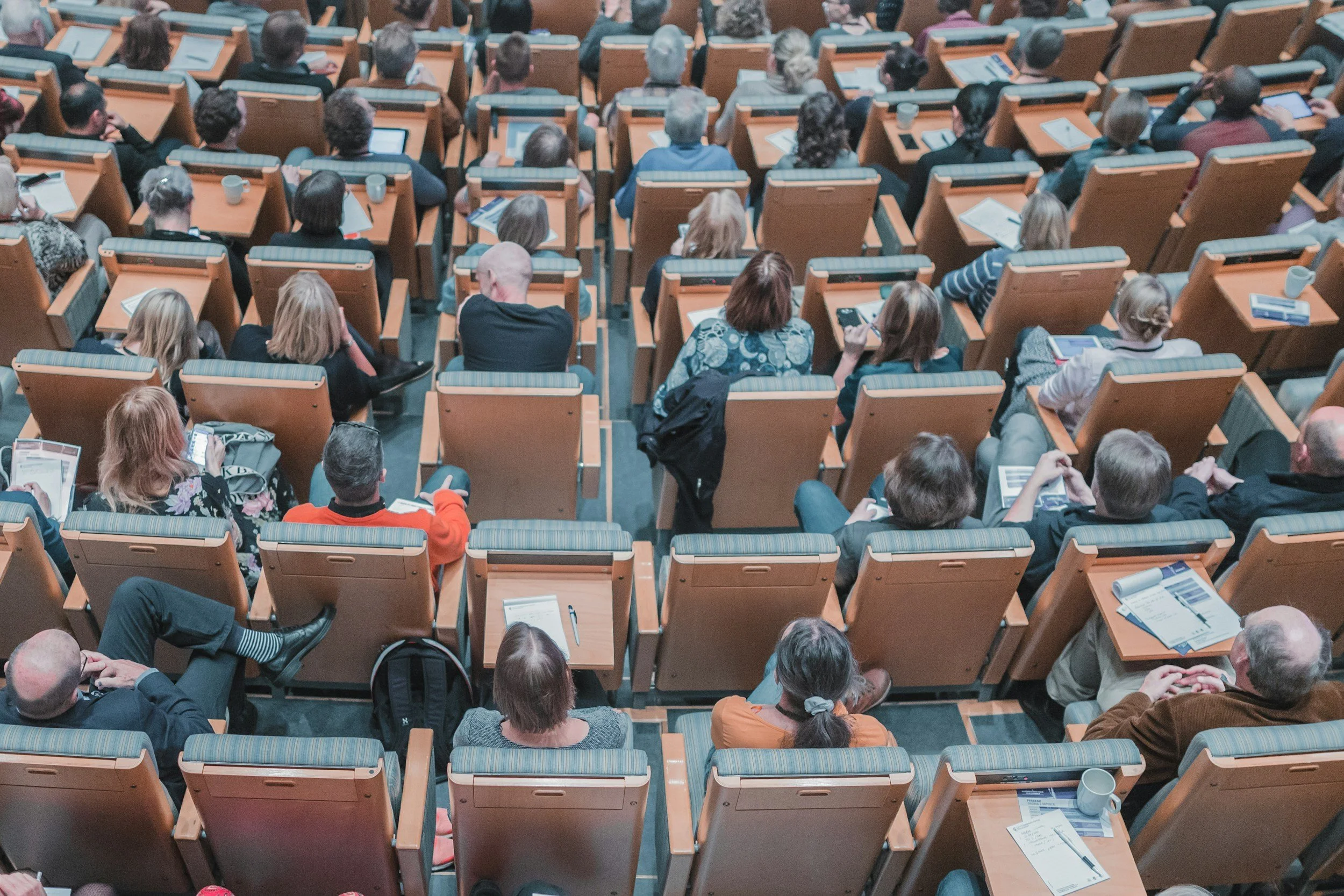 View of an auditorium filled with people sitting in wooden seats, attending a presentation or lecture, with many using tablets or notebooks.
