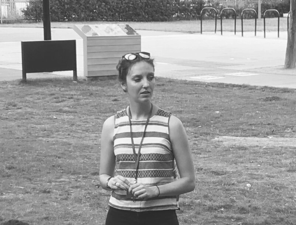 A woman standing outdoors in a park, wearing a striped sleeveless top, holding her hands together, with glasses resting on her head and trees and park equipment in the background.