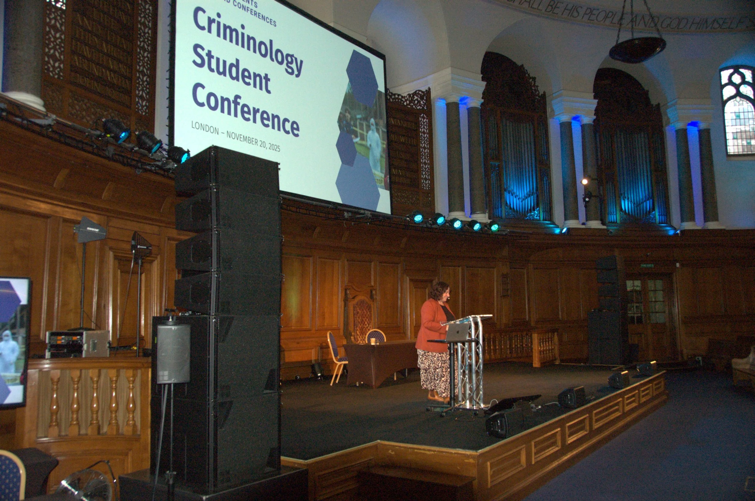 A woman speaking at a podium on a stage during the Crimeology Student Conference in London on November 20, 2025. There is a large digital screen displaying the conference name and date, with a wooden-paneled backdrop and blue lighting in the venue.