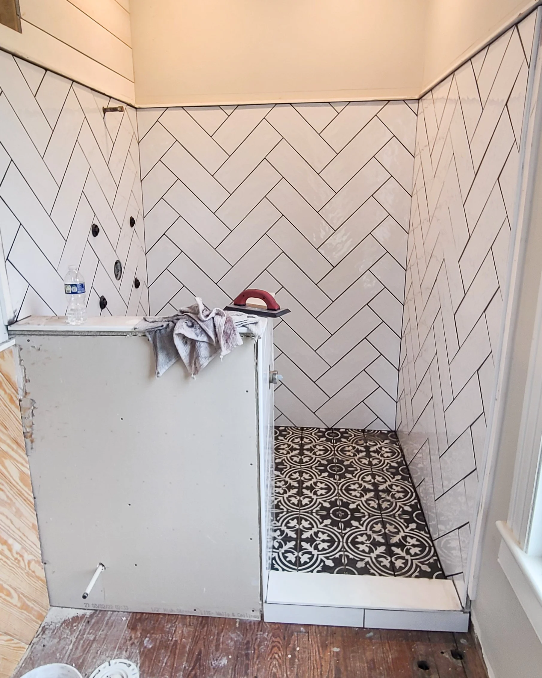 A bathroom under renovation with a partially installed shower, a towel, a water bottle, and a sander placed on the shower enclosure. The walls are decorated with white herringbone tiles, and the floor features black and white patterned tiles.