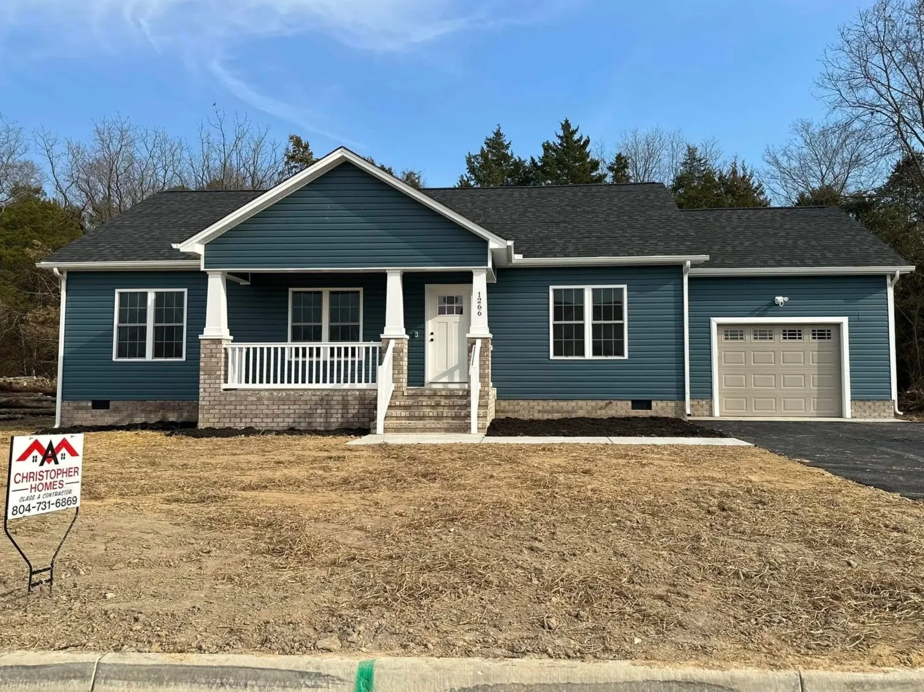 Newly built blue house with white trim, front porch, and attached garage, situated on a residential lot with a partially dirt yard, under a clear sky with sparse clouds.