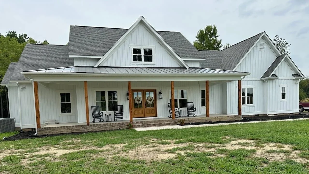 Front view of a white two-story house with a covered porch, wooden support beams, and a brick foundation, set on a grassy lot.