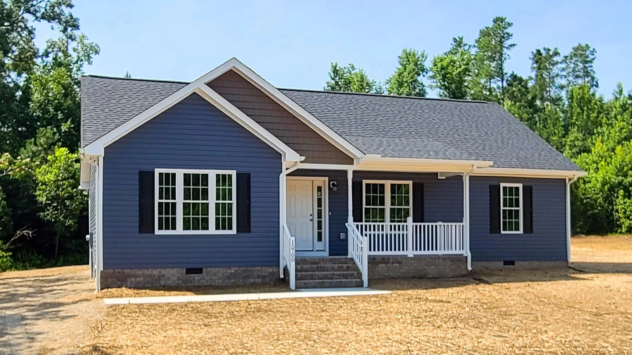 Blue house with white trim, black shutters, front porch with white railing, set on a dirt yard, surrounded by green trees under a clear blue sky.