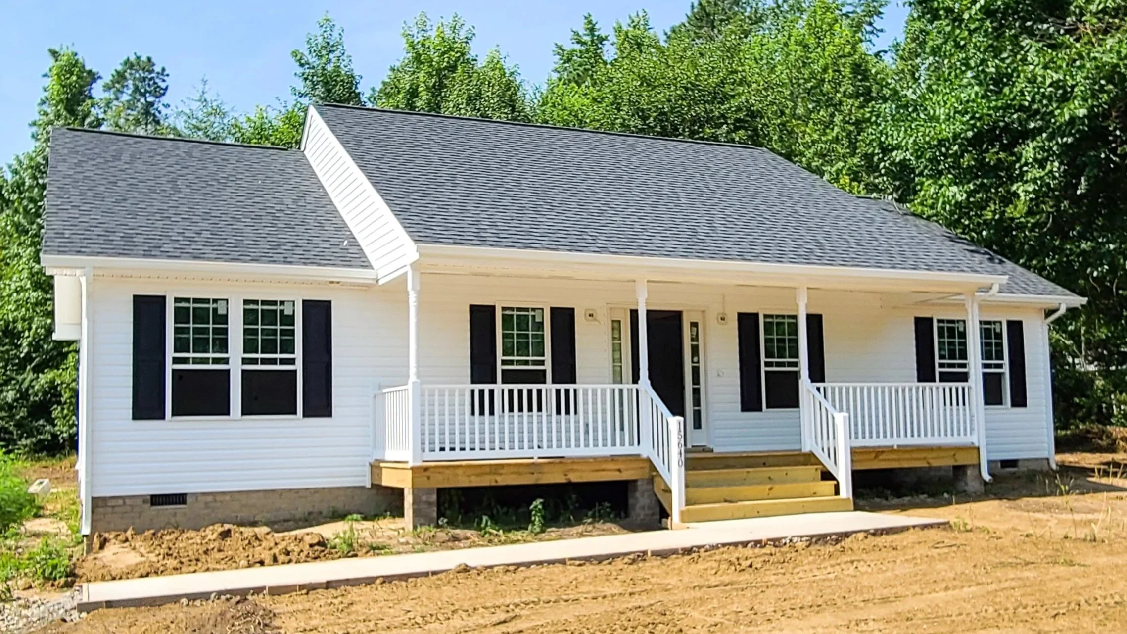 New white house with black shutters and a gray roof, with a front porch and steps, surrounded by trees and dirt ground.