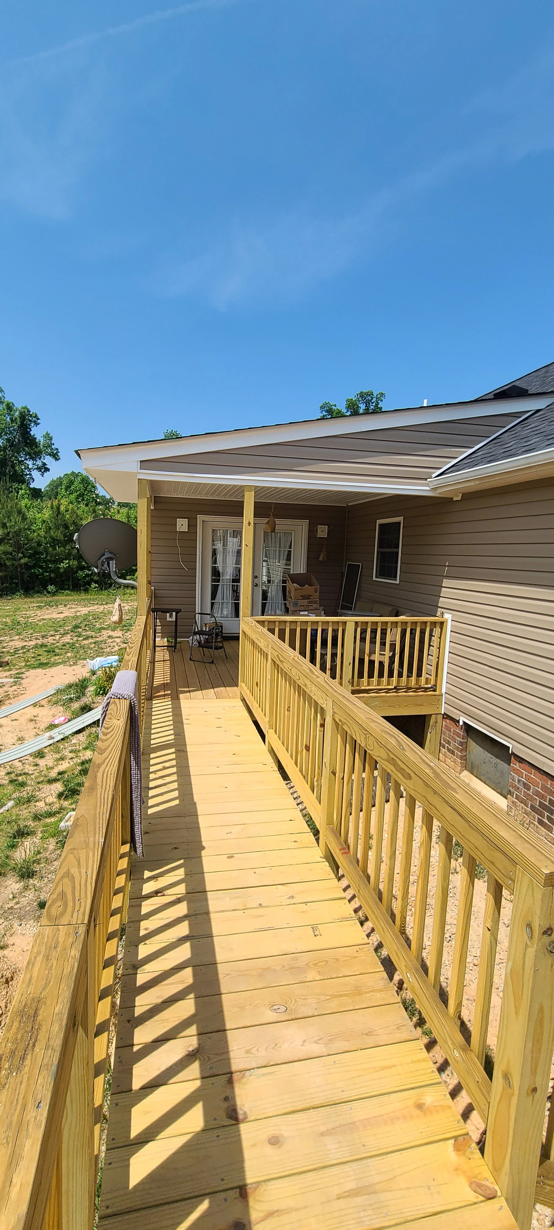 New wooden deck attached to beige house with small staircase, sliding glass door, and satellite dish outside under clear blue sky.